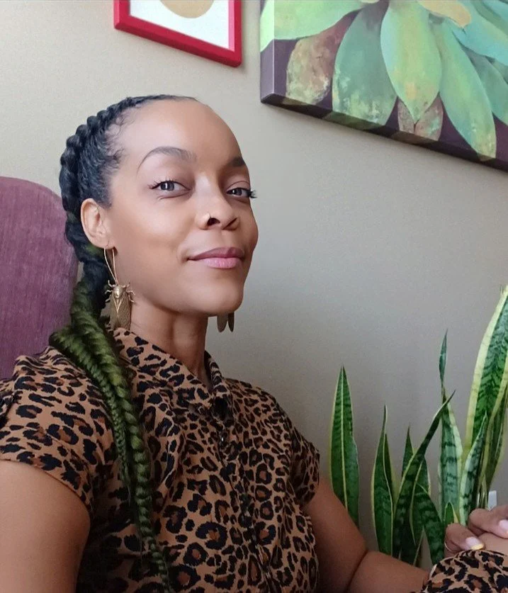 A woman with braided green hair wearing a leopard print shirt, sitting in a room with a plant beside her and framed artwork on the wall behind her.
