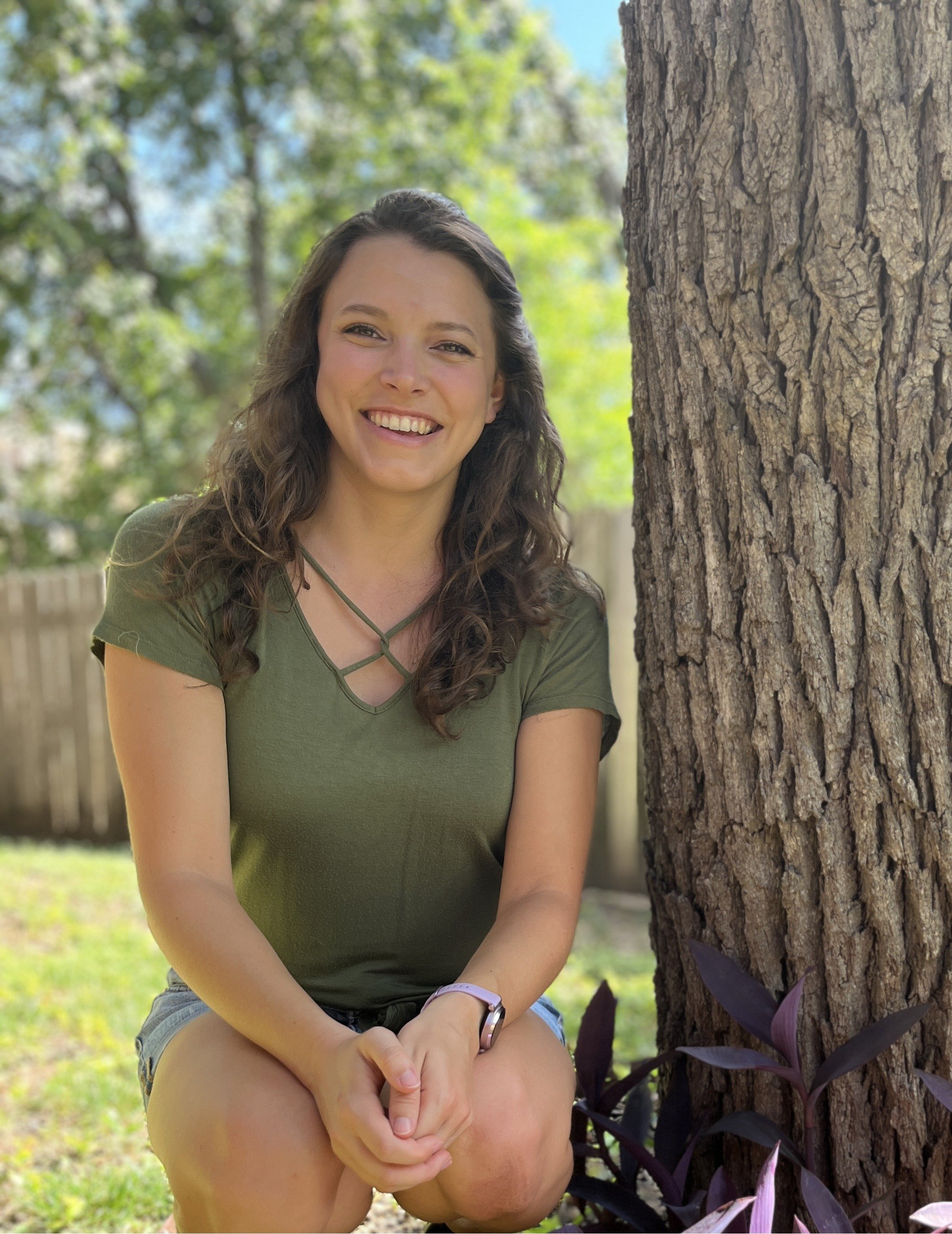 A woman with long curly brown hair smiling, wearing a green top and shorts, sitting outdoors beside a large tree trunk with green plants and a wooden fence in the background.