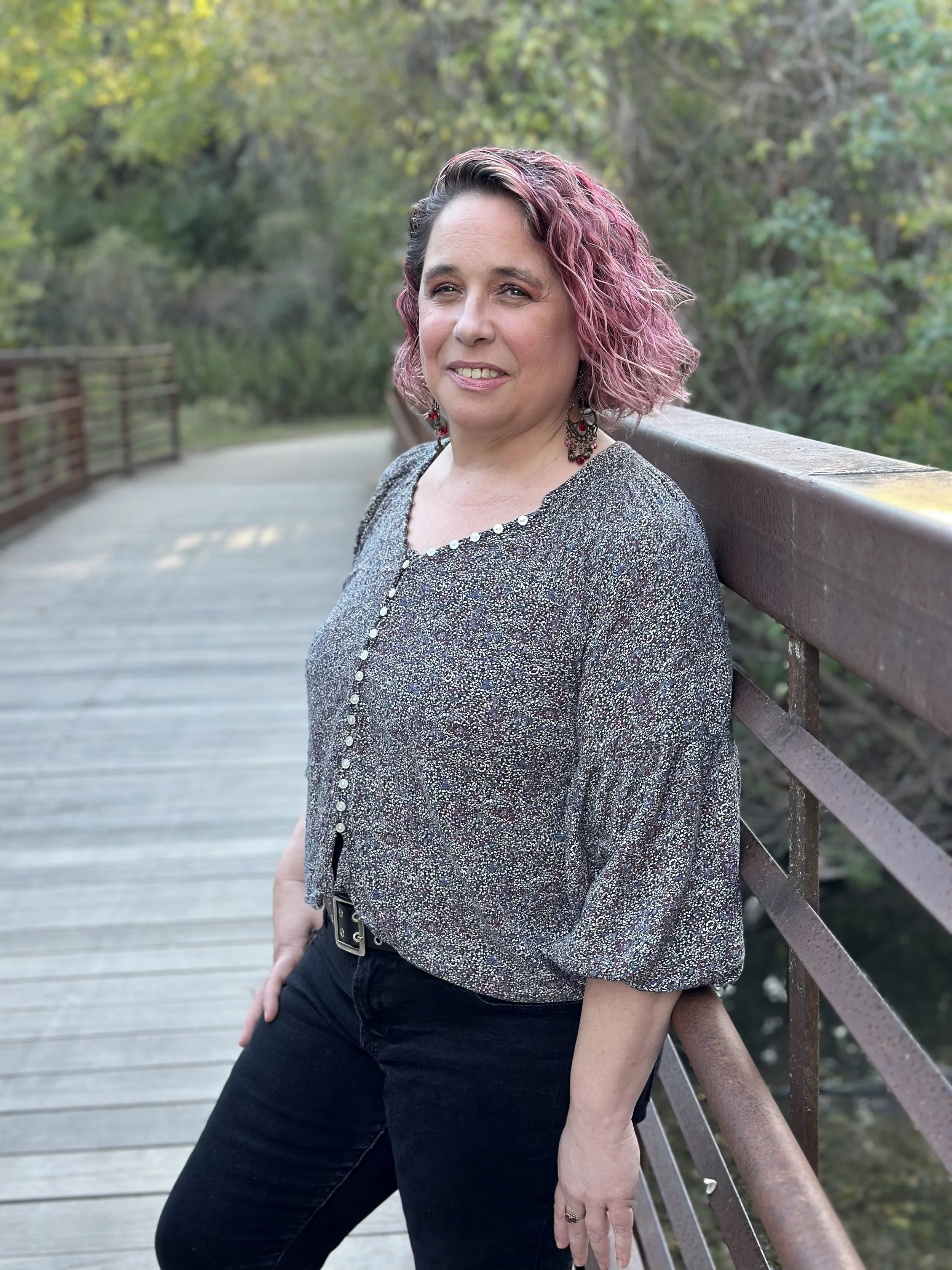 A woman with pink, curly, shoulder-length hair stands on a wooden bridge outdoors, leaning against the railing and looking at the camera with a slight smile. She is wearing a shiny, patterned blouse and black pants.