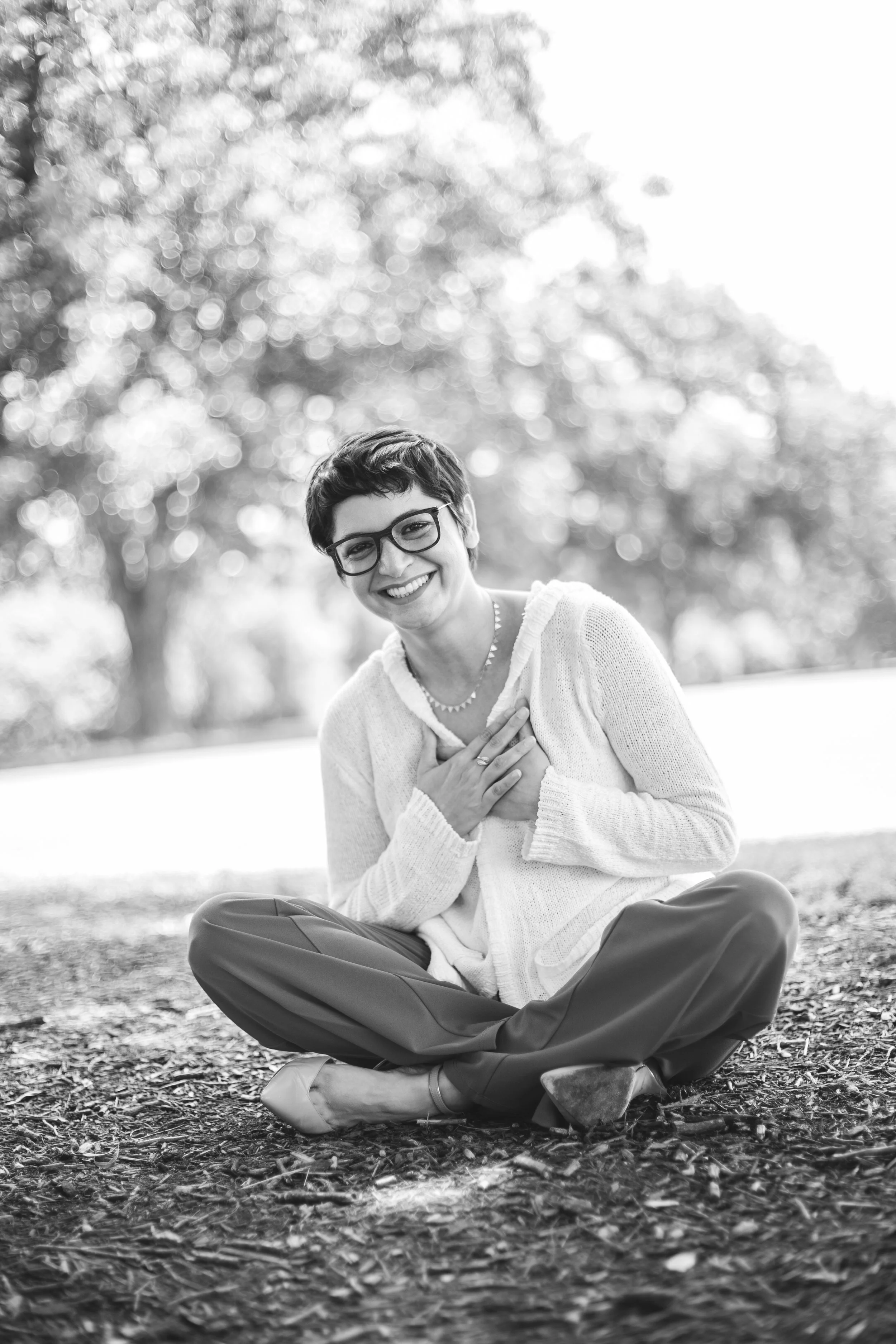 A woman sitting cross-legged on the ground outdoors, smiling and holding her hand over her chest, in a park with trees in the background.