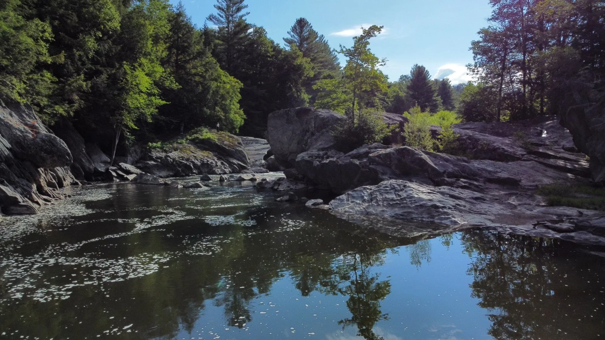 A river flowing through a rocky landscape lined with green trees under a blue sky.