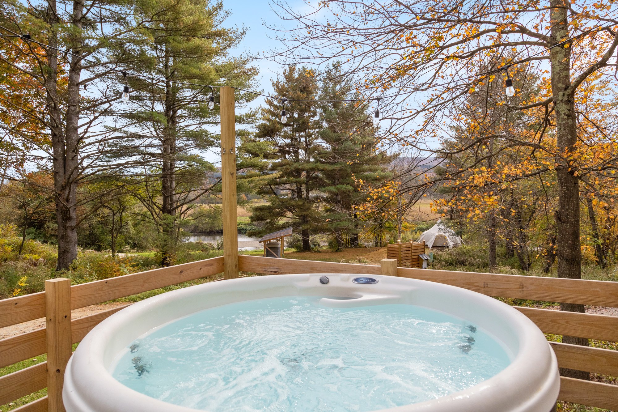 Outdoor hot tub on a wooden deck surrounded by trees with autumn leaves, string lights hanging above, overlooking a river and landscape in the distance.