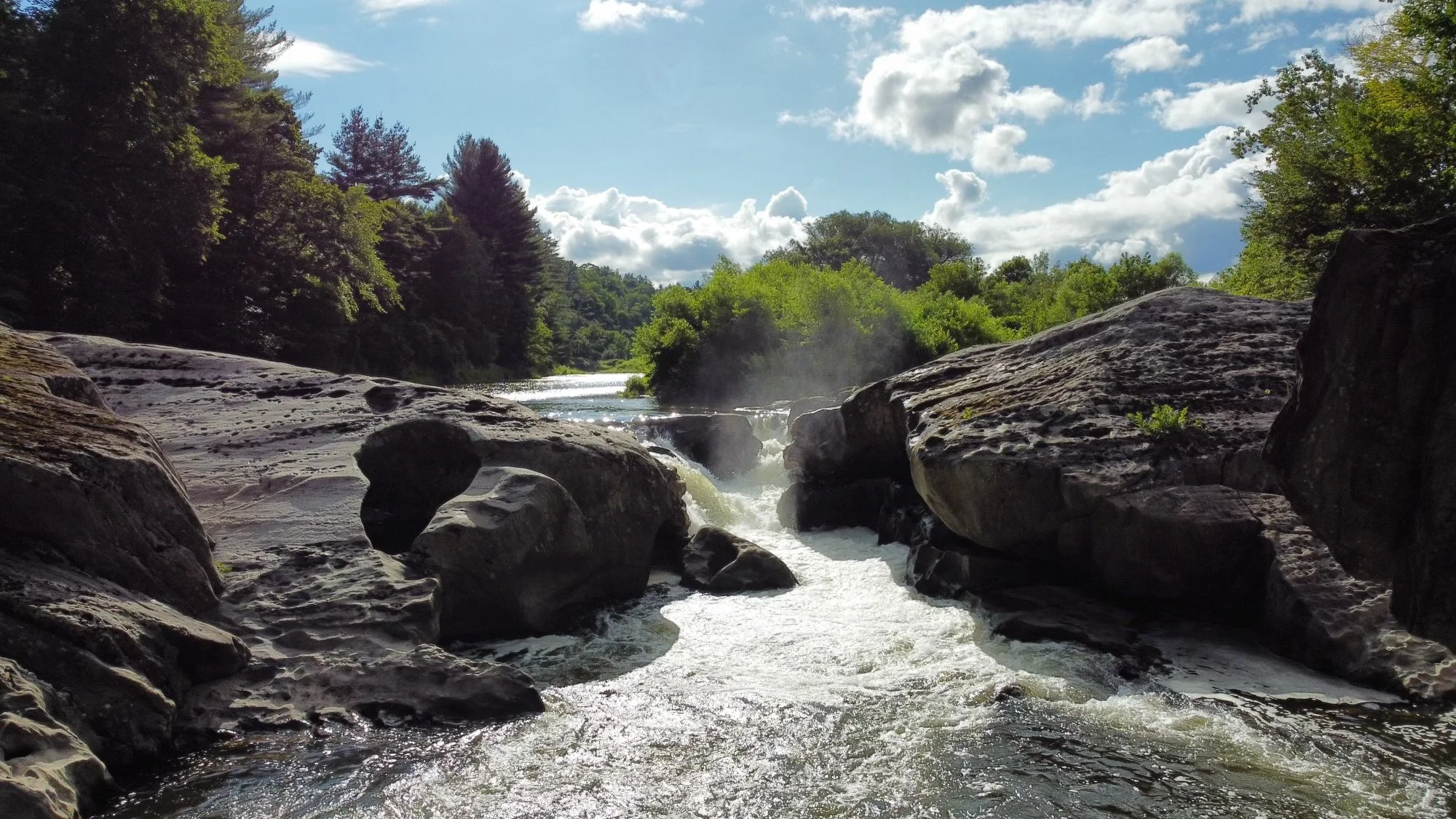 A river cascading over rocks surrounded by trees on a sunny day with partly cloudy sky.