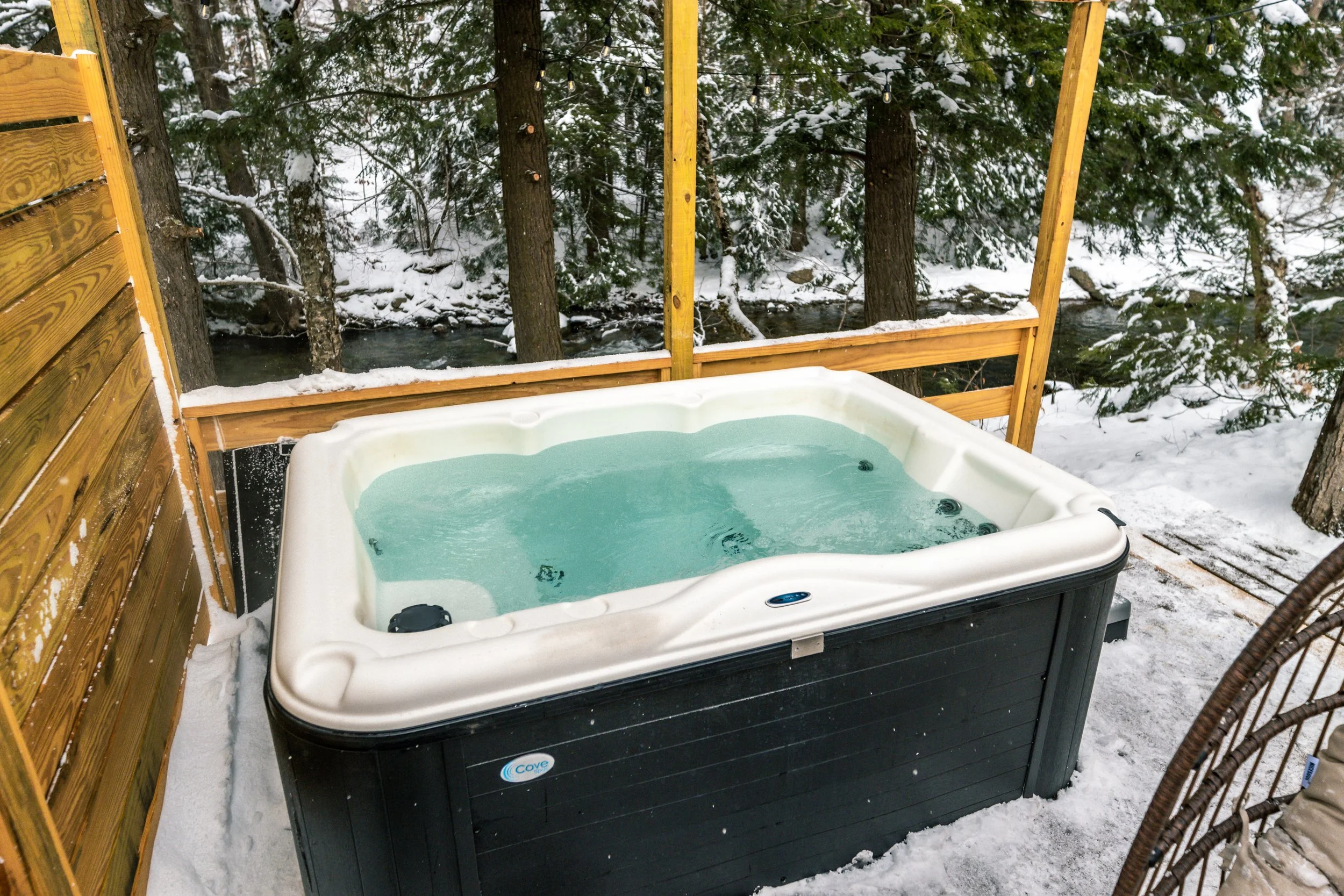 Outdoor hot tub with clear water on a snow-covered deck, surrounded by trees with snow, overlooking a river in a winter forest setting.