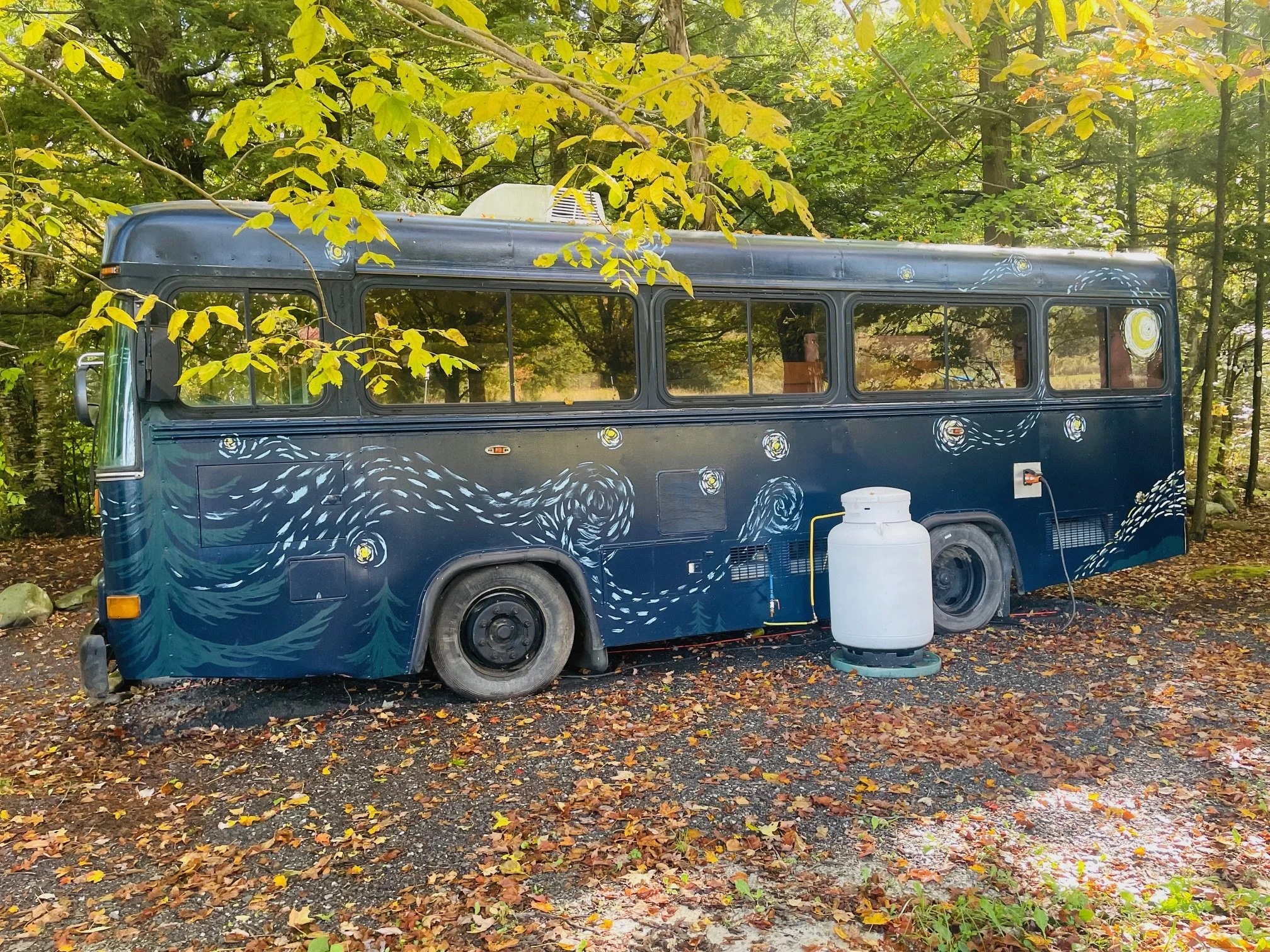 A camper van painted with a starry night pattern, parked on a wooded area with fallen autumn leaves on the ground. There is a white propane tank beside the van.