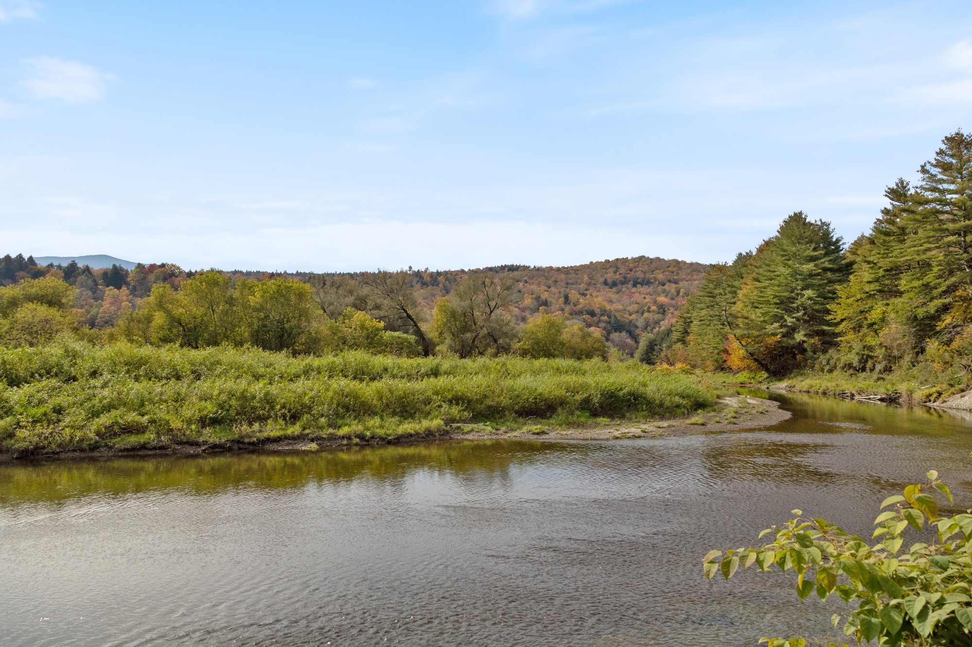 A river flowing through a lush landscape of trees with fall foliage, under a partly cloudy sky.