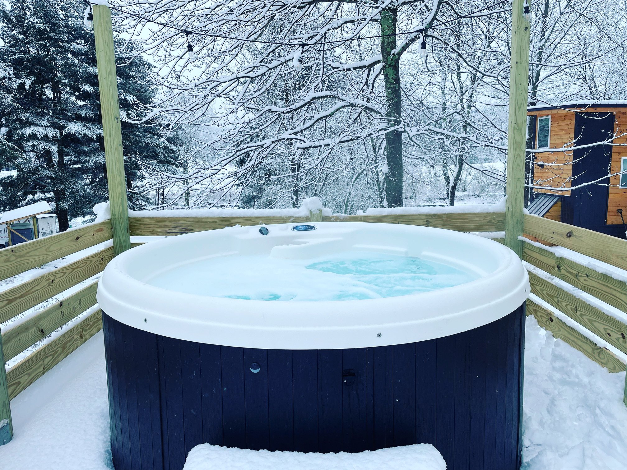 An outdoor hot tub on a wooden deck with snow, surrounded by snow-covered trees and a wooden house in the background.