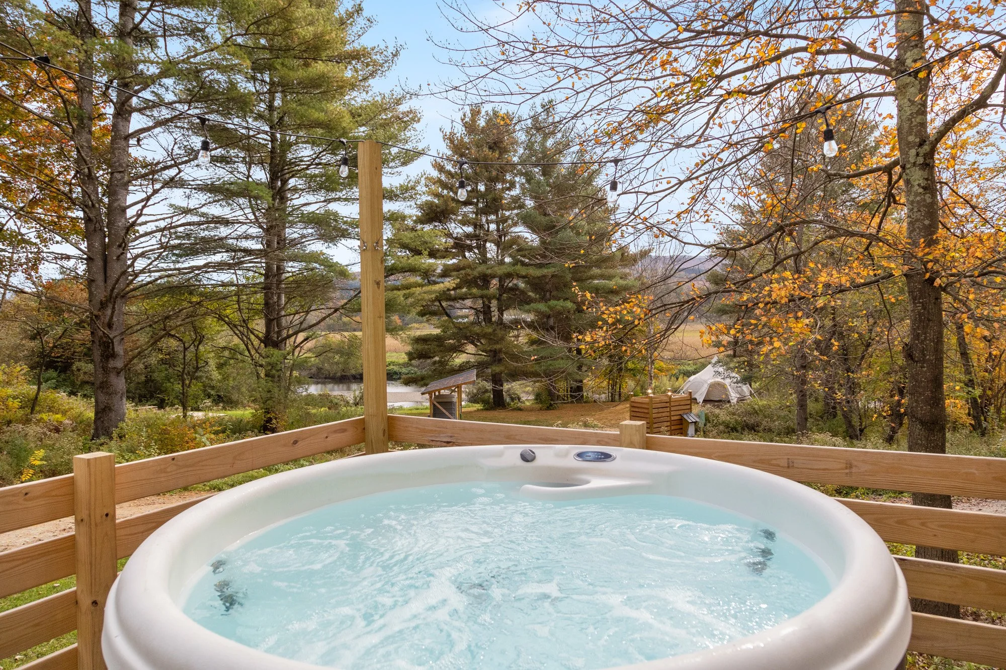 Hot tub on a wooden deck overlooking trees and a river in a wooded area during fall with orange and yellow leaves.