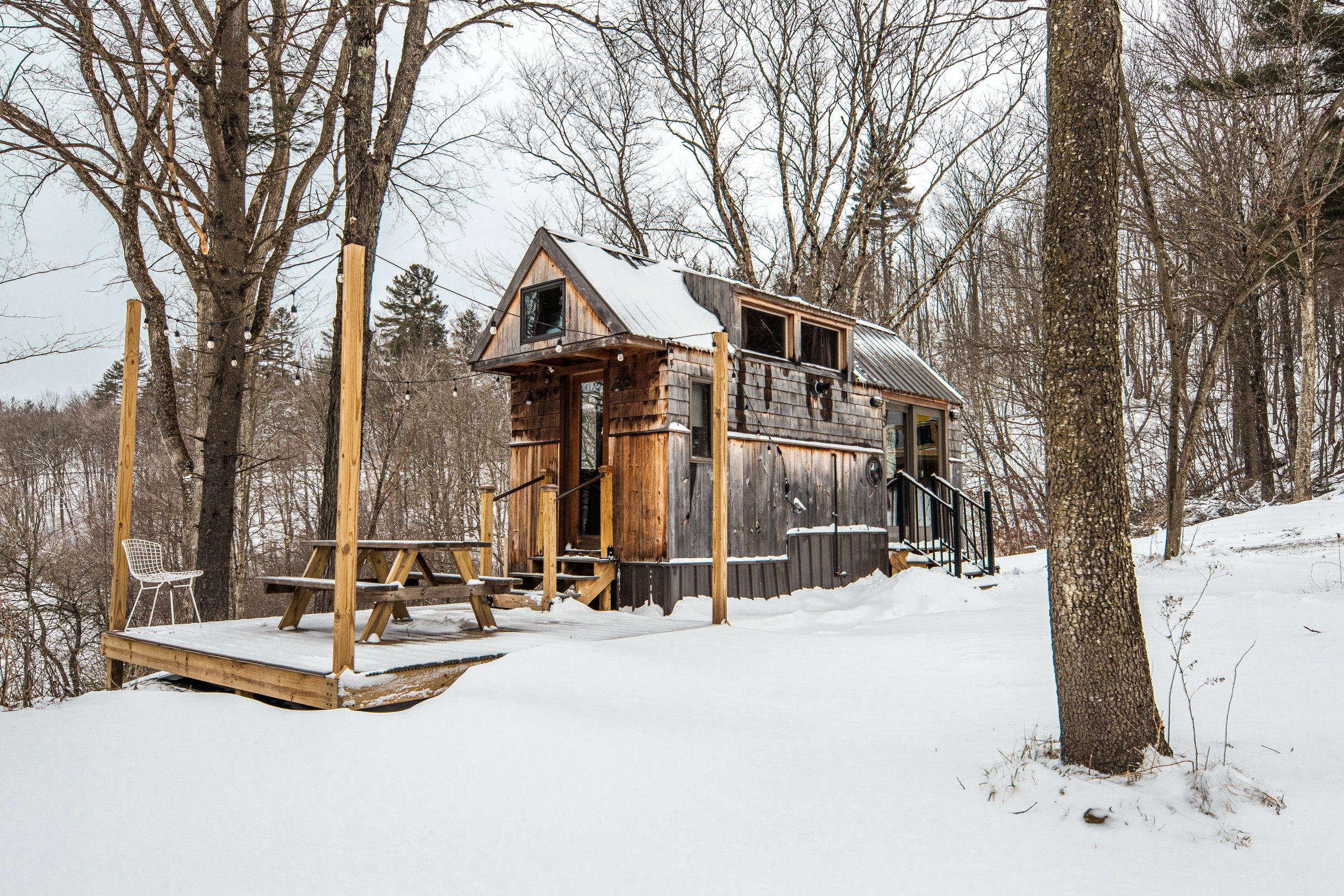 Small wooden cabin in a snowy landscape with bare trees, a deck with a picnic table and a chair, and string lights hanging above.