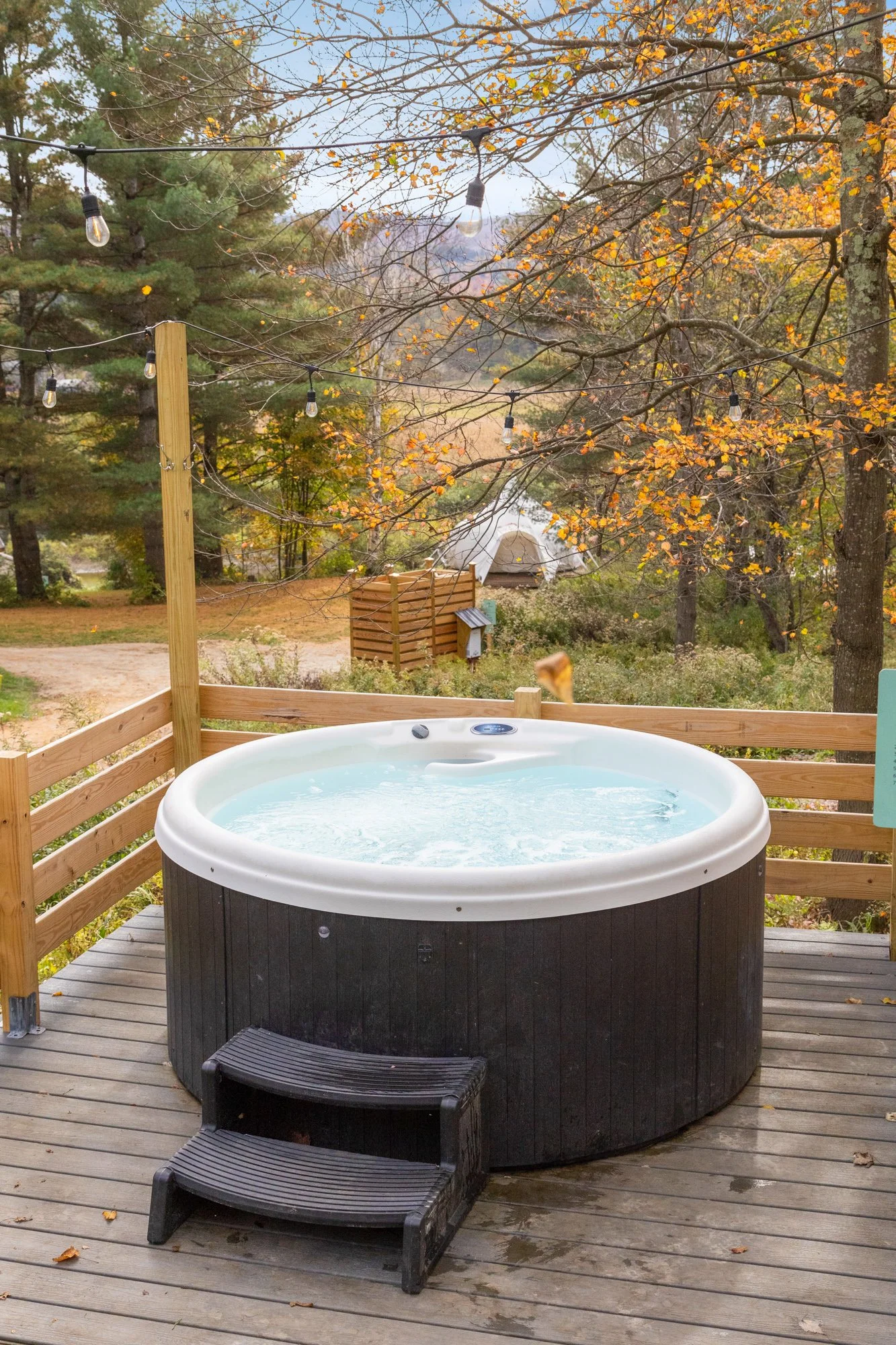 An outdoor hot tub on a wooden deck with a scenic autumnal background, string lights overhead, and a tent in the distance.
