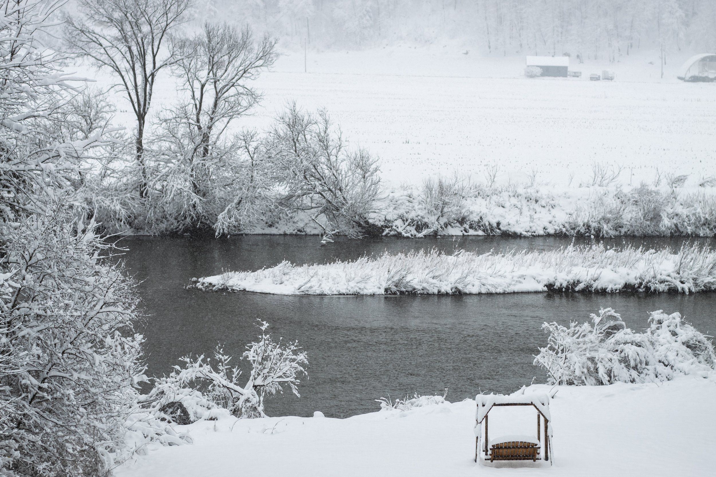 Snow-covered landscape with a river, snow-laden trees, and a wooden swing in the foreground.