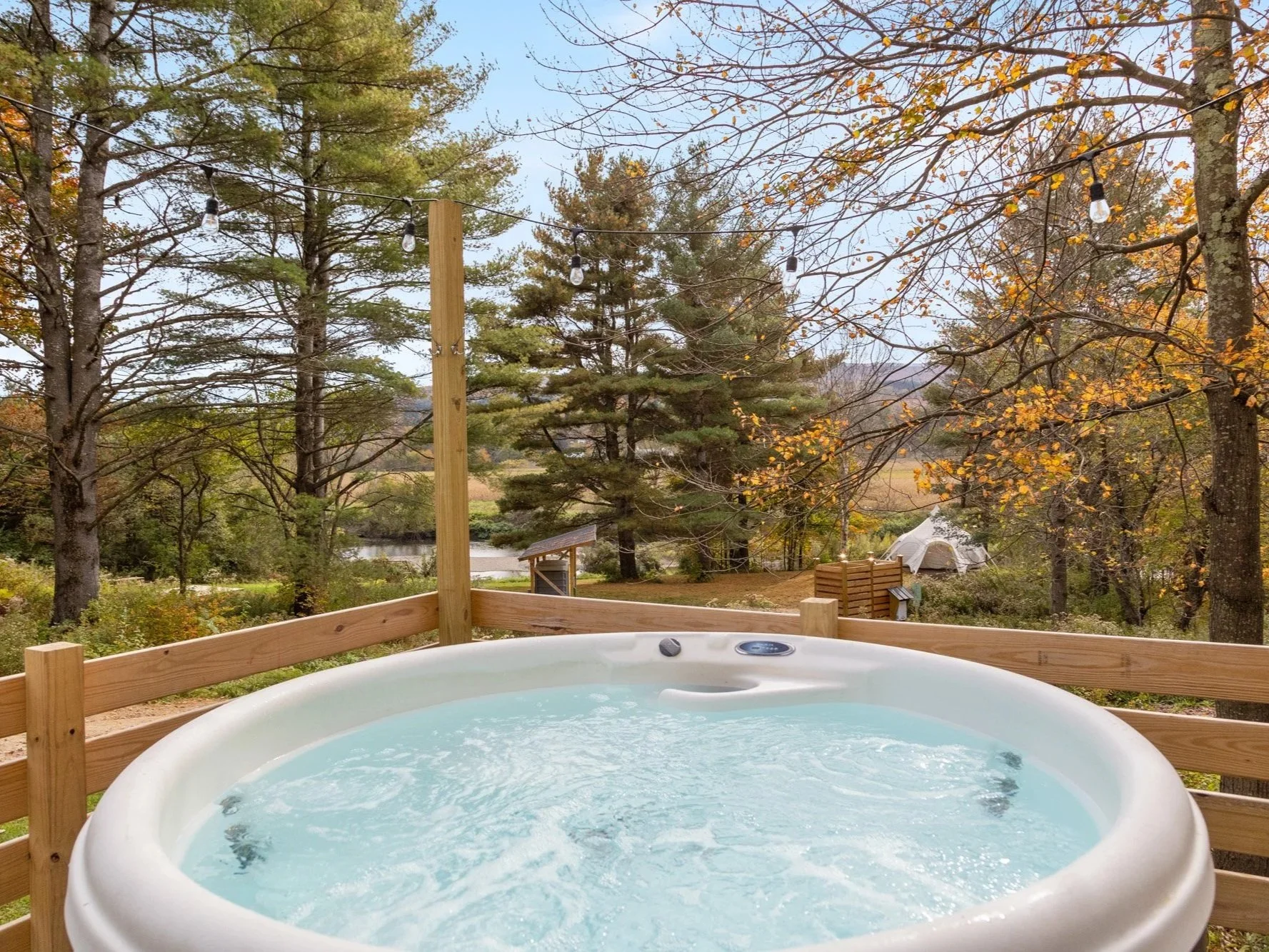 An outdoor hot tub on a wooden deck surrounded by trees with autumn foliage, with string lights overhead and a view of a lake and tent in the background.
