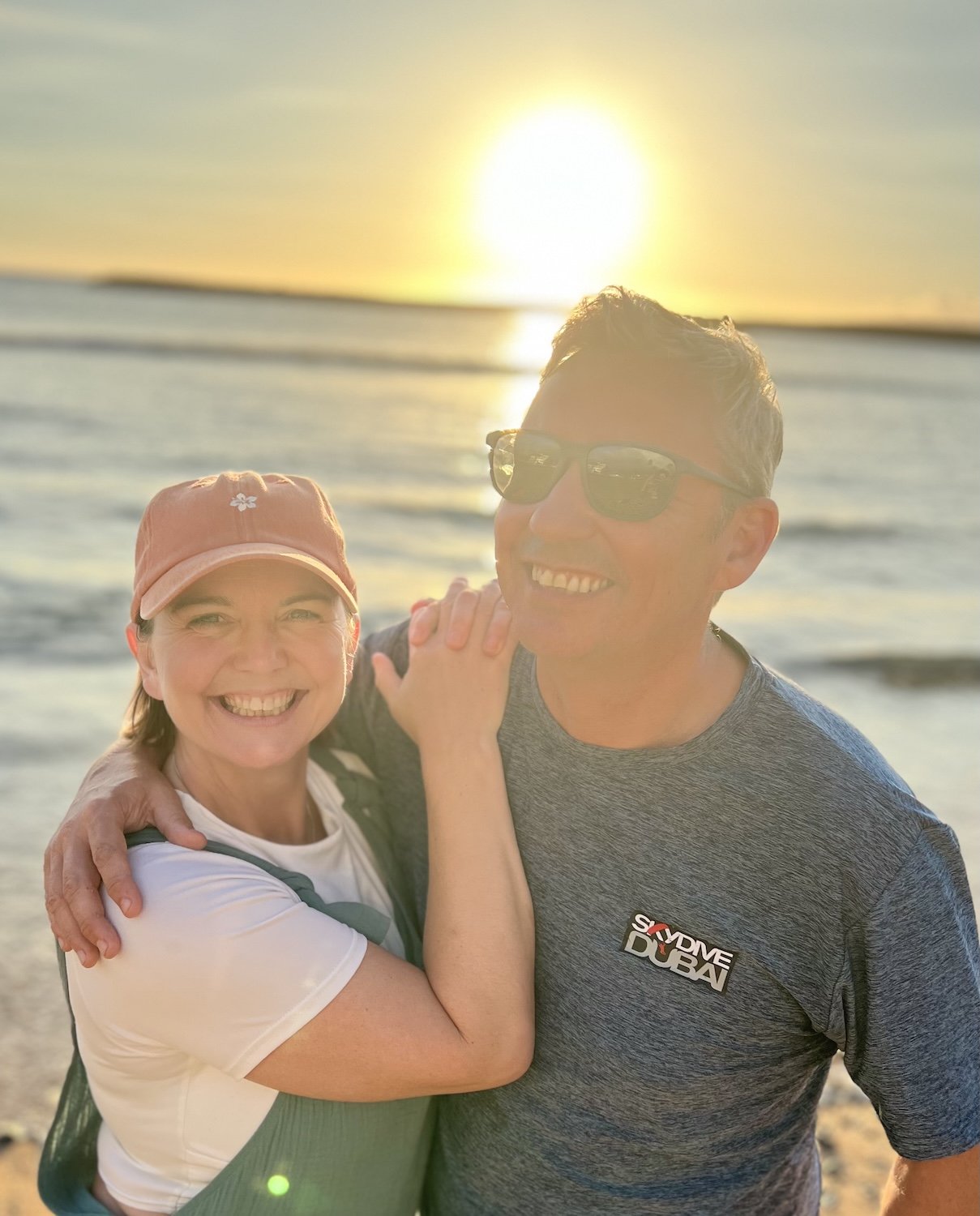 Shona and Paul Morgan smiling with a view of the sea and a sunset behind them - TXM Campers