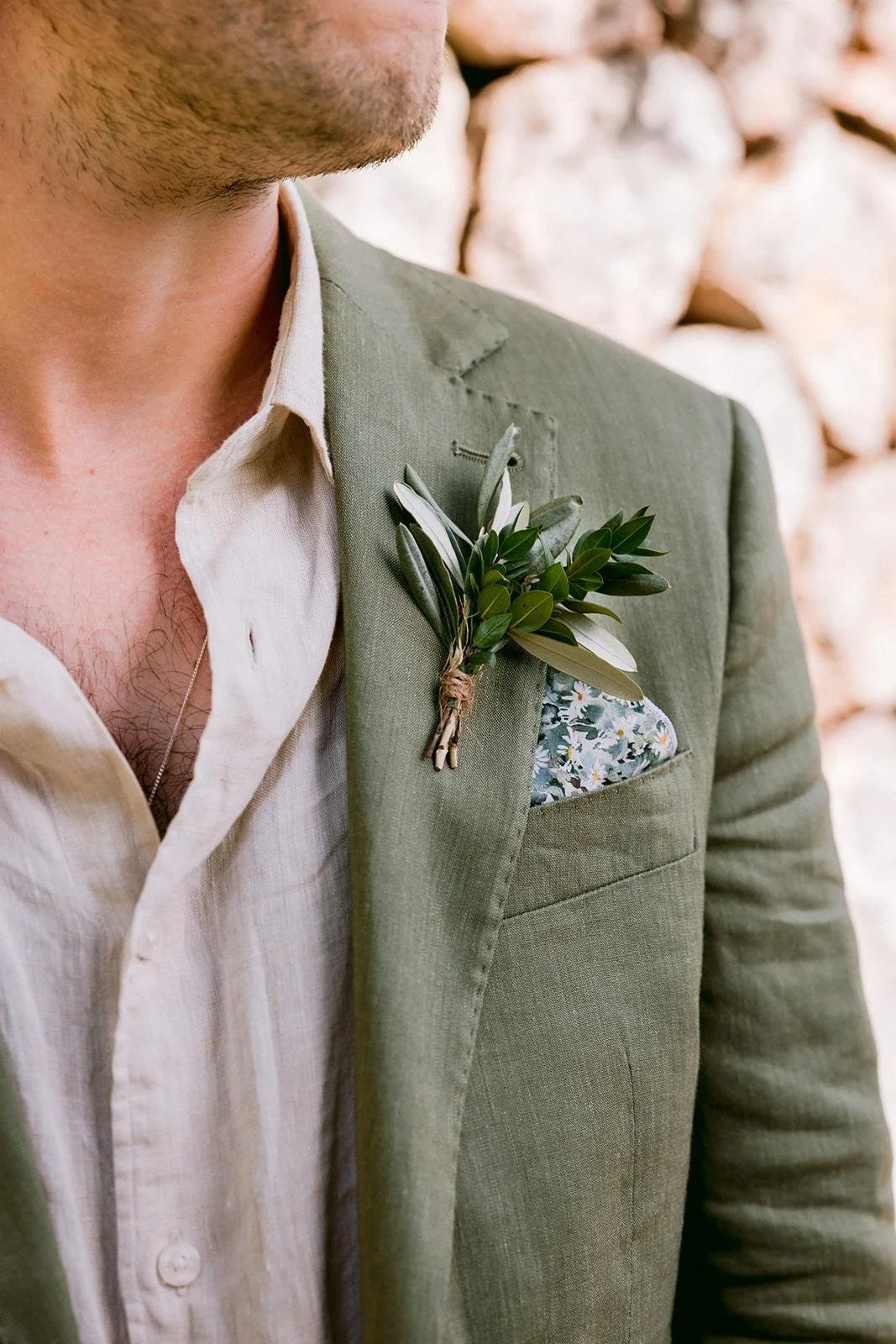 Close-up of a man wearing a light green suit jacket with a floral pocket square and a boutonniere made of greenery, against a background of stacked stones.