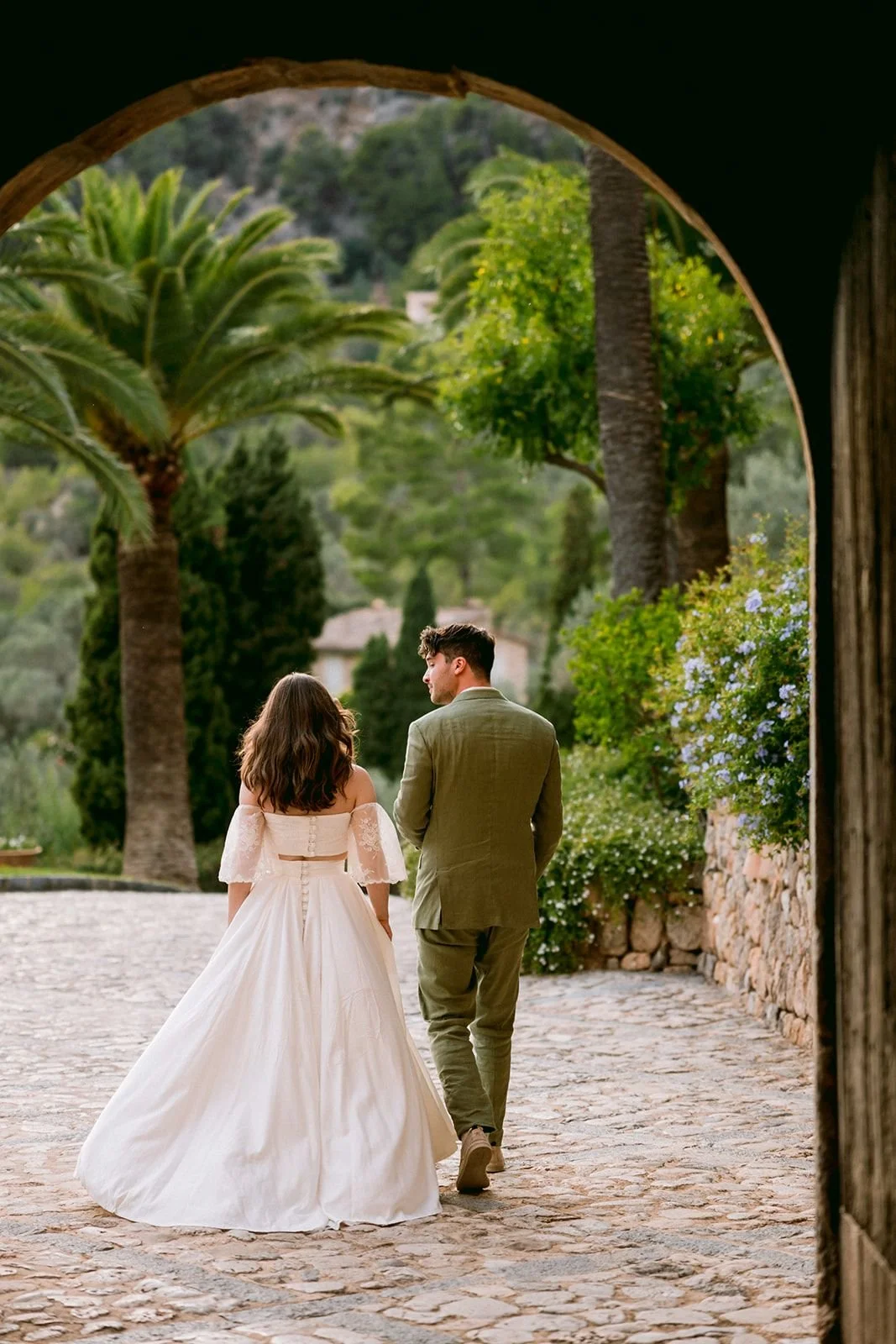 Couple walking on stone pathway outdoors, framed by an archway, with lush green trees and plants in the background.