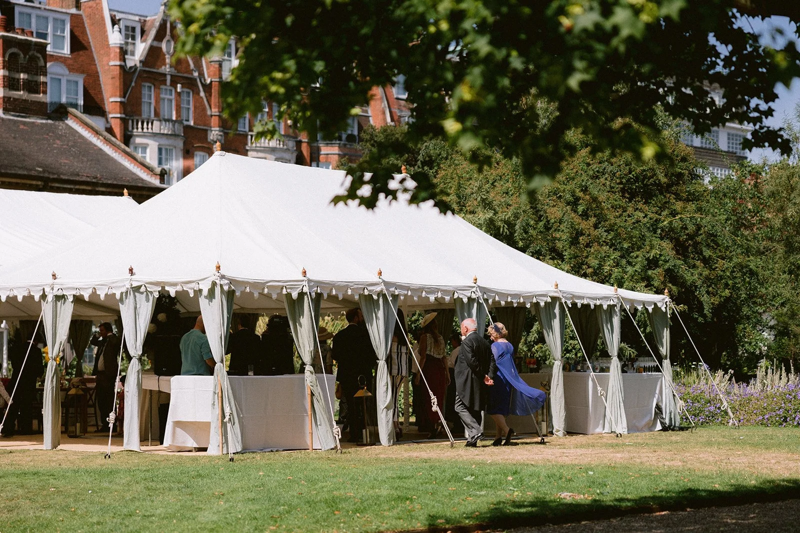 A large white event tent set up outdoors on a lush green lawn with guests inside and outside, surrounded by trees and a historic brick building in the background.