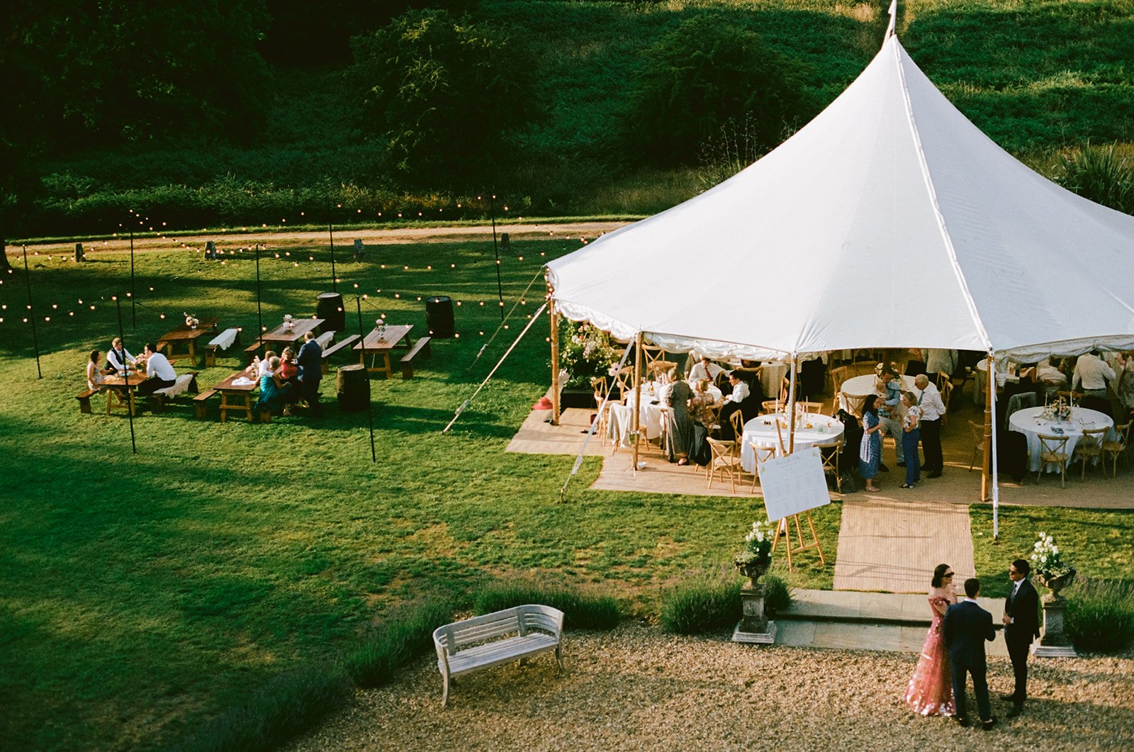 Outdoor wedding reception with a large white tent, decorated tables, and guests mingling on a lawn with string lights and a garden.
