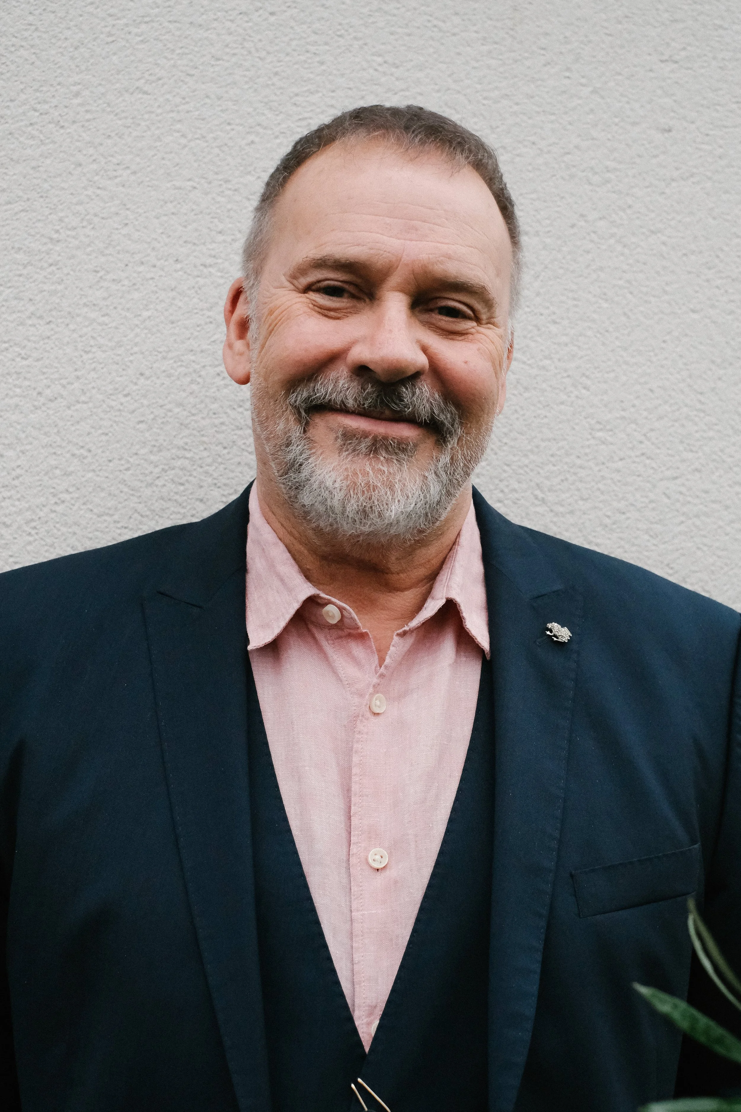 A middle-aged man with a beard and mustache, smiling, wearing a pink shirt and a dark blazer, standing against a plain wall.