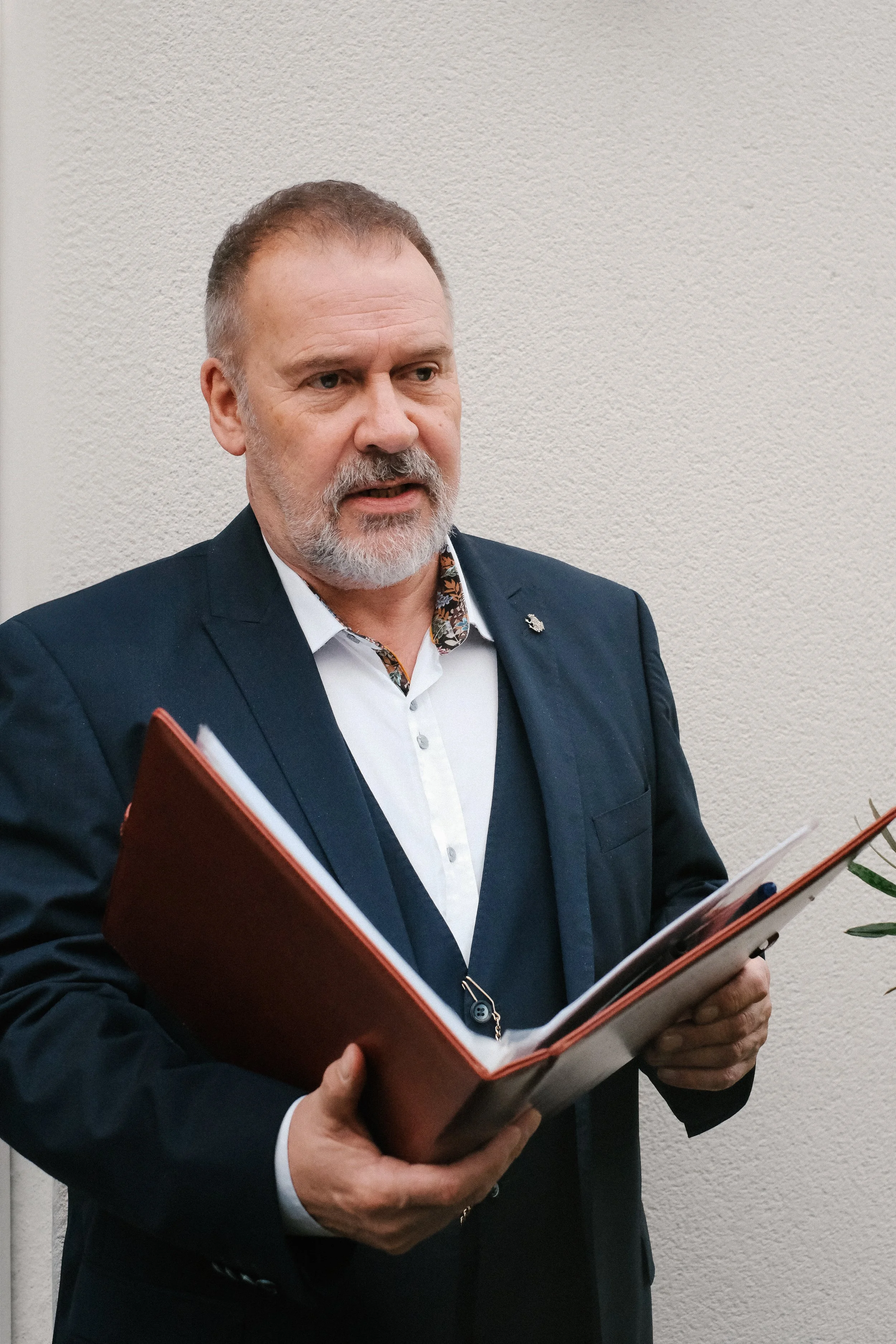 A middle-aged man with gray hair and a beard is wearing a dark suit jacket, a white shirt, and a vest. He is holding an open folder or binder and appears to be speaking.
