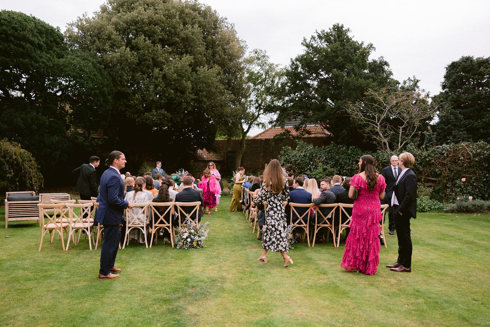 An outdoor wedding ceremony with guests seated on wooden chairs on a green lawn, surrounded by trees and bushes, with the officiant and bride standing at the altar.