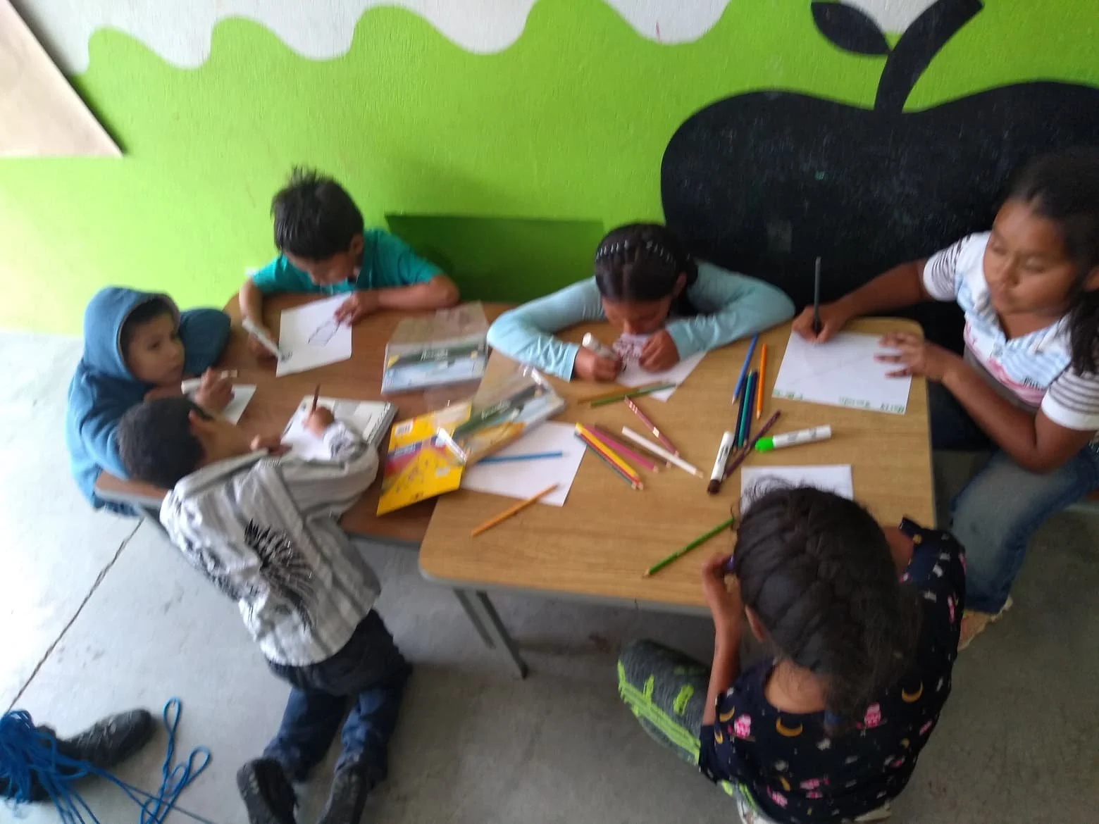 Children and an adult sitting and drawing at a table, with various colored pencils and paper, in a room with light-colored tile floor and a bright green wall with black apple-shaped design.