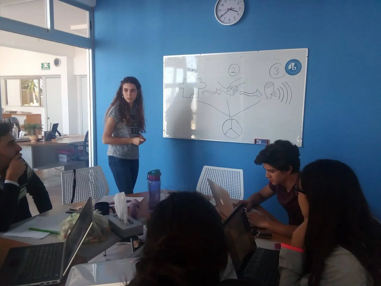 Young woman giving presentation to a group of students in a classroom with a whiteboard and laptops.