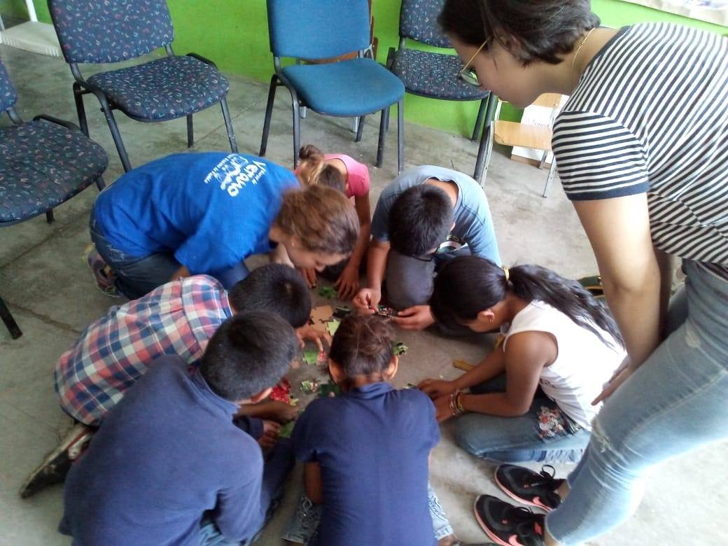 Group of children and a woman sitting on the floor, working on a puzzle together indoors with bookshelves and chairs in the background.