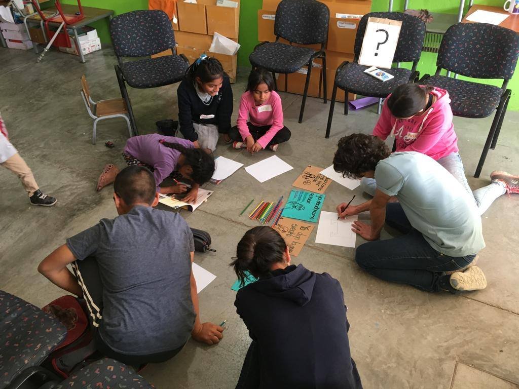 A group of children and an adult sitting on the floor in a classroom, working on papers and colored pencils, with chairs, boxes, and a green wall with a question mark poster in the background.