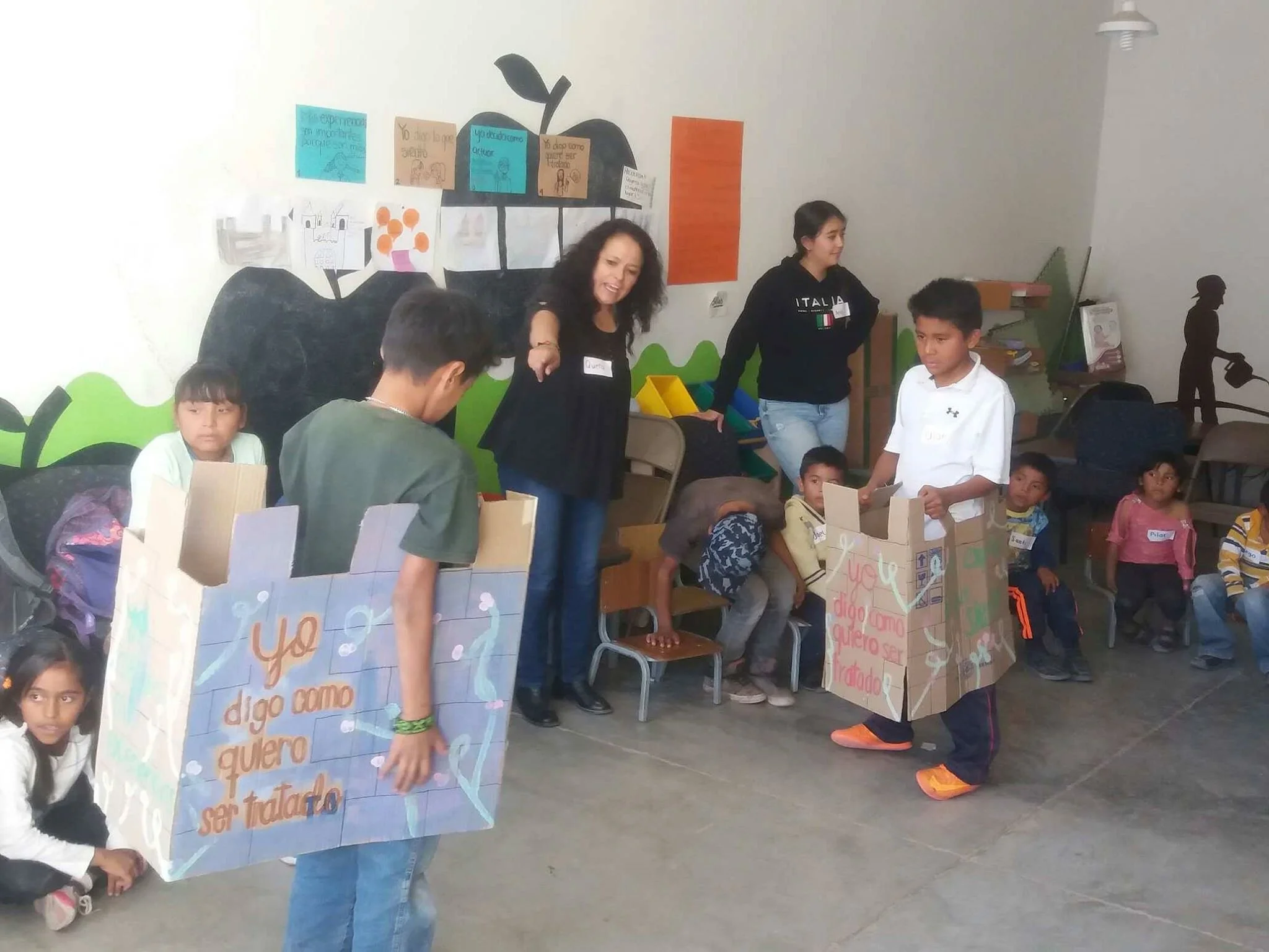 Children and adults participating in a classroom activity, holding large cardboard signs with handwritten messages in Spanish. The classroom has colorful decorations and a wall mural with a tree and posters.