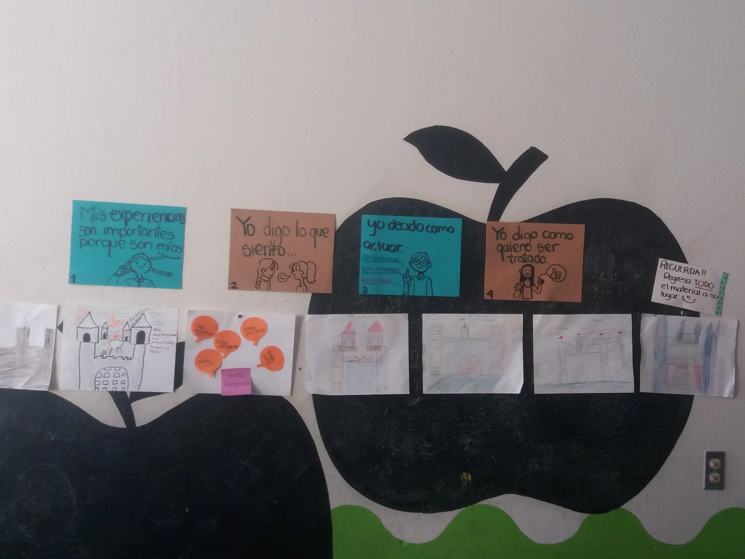 School bulletin board decorated with paper castle drawings, sticky notes, and handwritten messages about experiences and feelings, mounted on a wall painted with a large apple design.