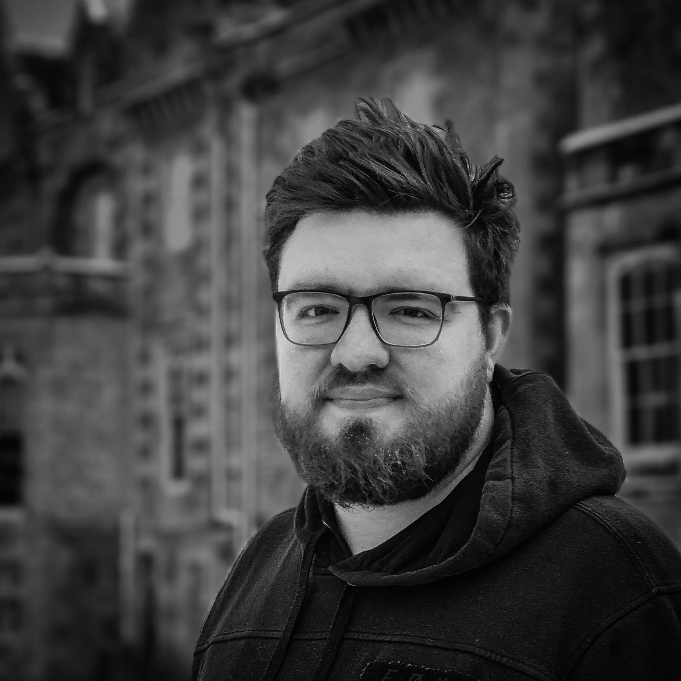 A man with glasses and a beard looks at the camera, standing outdoors in front of an old brick building with arched windows.
