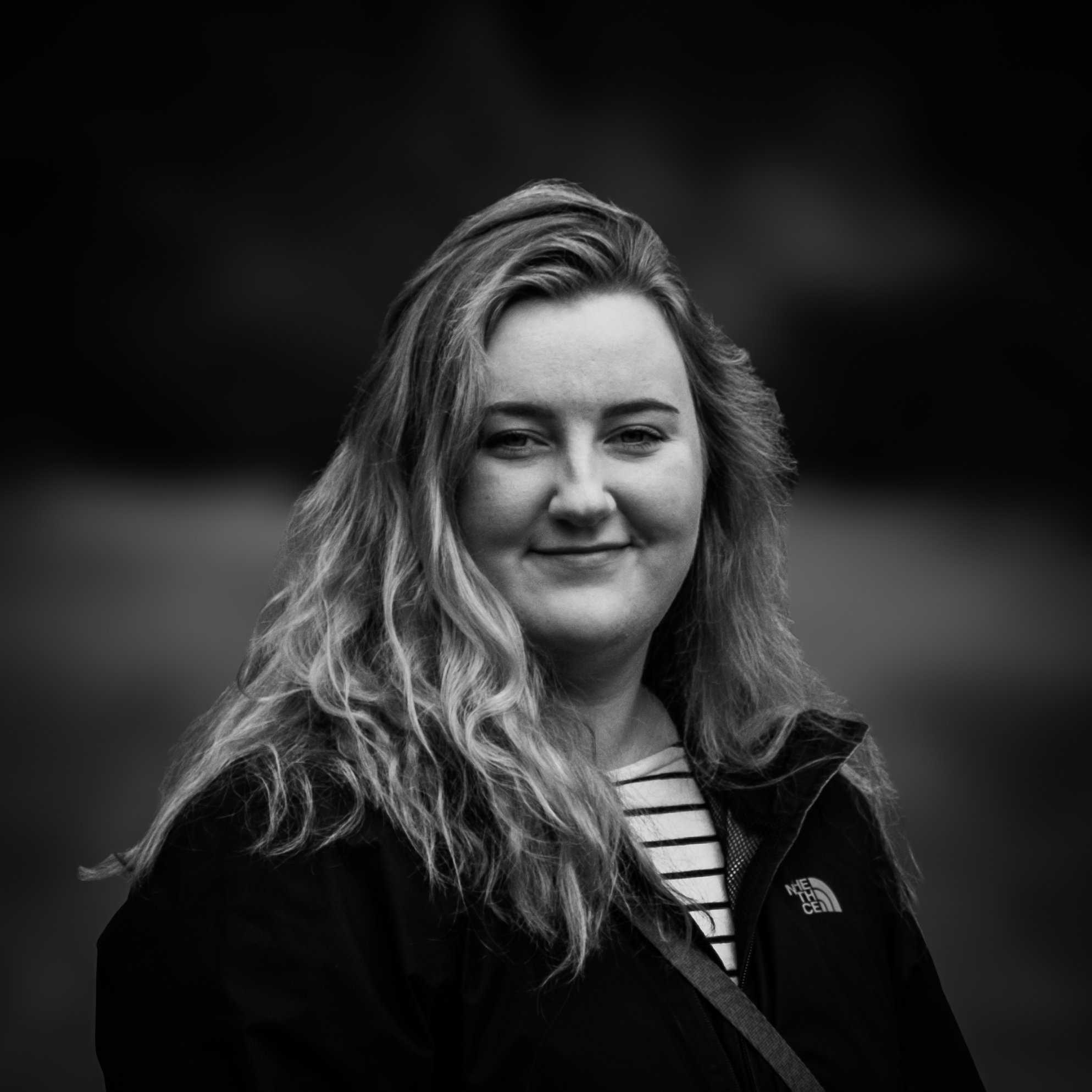 Black and white portrait of a woman with wavy hair smiling, wearing a North Face jacket and striped shirt, outdoors with a blurred background.