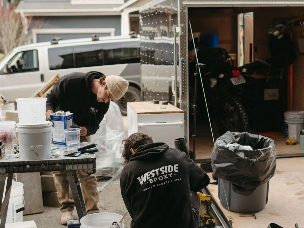 Two people working for Westside Epoxy on a garage floor, with buckets, tools, and supplies around them. One person is bending, the other is sitting with their back to the camera about to mix epoxy for the floor.