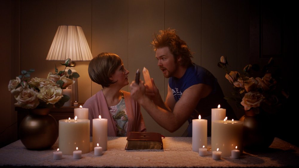 Jennifer in a pink cardigan at a candlelit table while a man waves a crystal at her