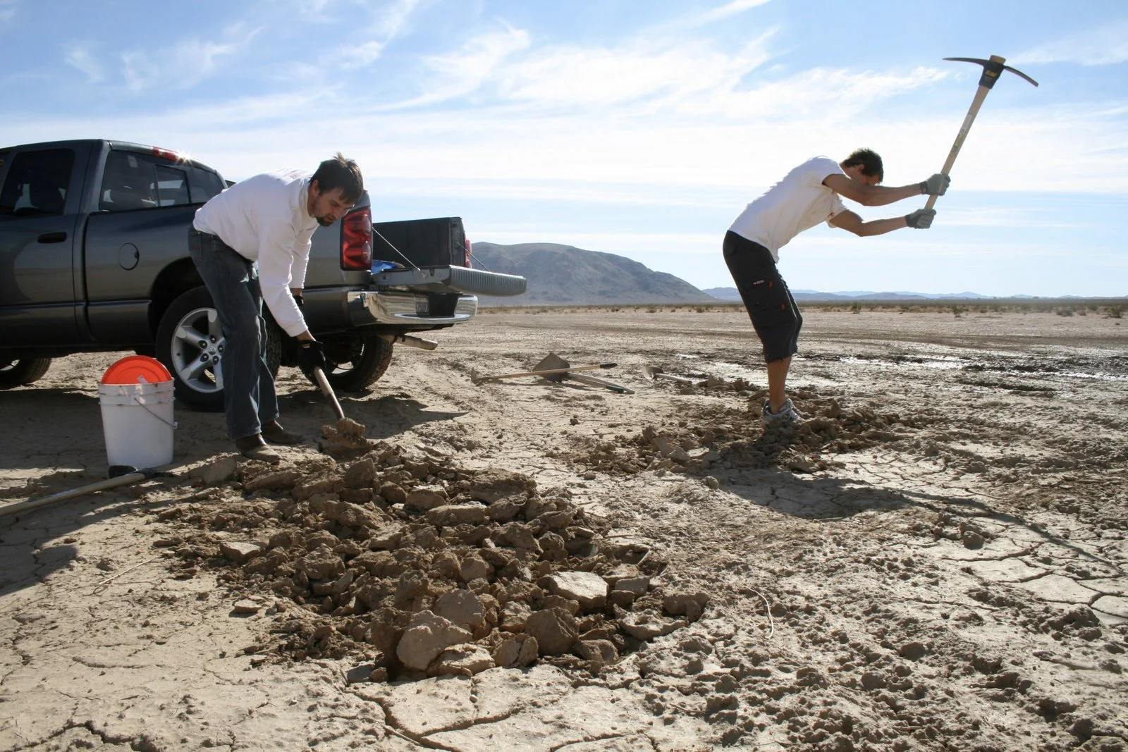 Two people, a man and a woman, working on a dry, cracked landscape near a black pickup truck. The man is using a shovel to dig into the ground, while the woman is wielding a pickaxe. There are tools and a bucket nearby, with mountains in the background under a blue sky.