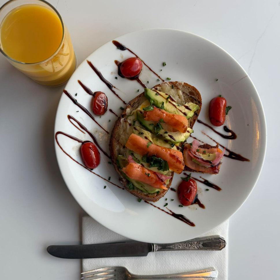 An open-faced sandwich topped with avocado, smoked salmon, and herbs on a slice of bread, drizzled with balsamic glaze. The plate is garnished with cherry tomatoes and balsamic drizzle. There is a glass of orange juice beside the plate.