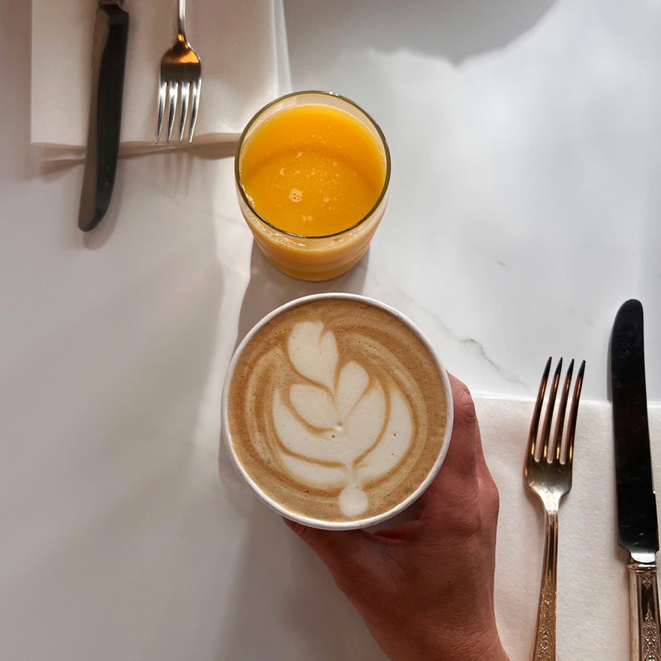 A hand holding a latte with latte art, a glass of orange juice, and a set of cutlery on a white table.