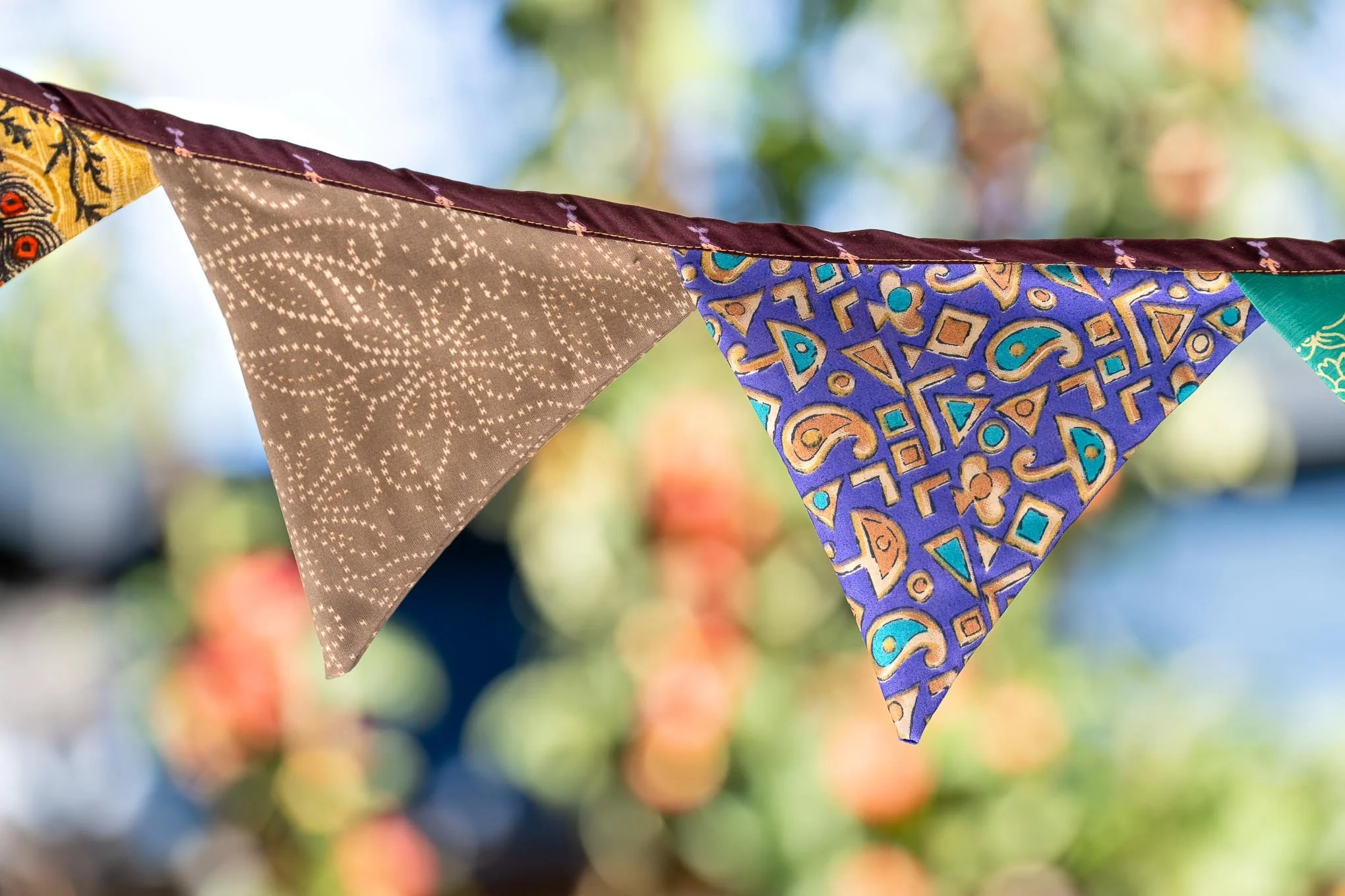 Bunte Stoffflaggen hängen an einer Schnur im Freien, im Hintergrund sind unscharfe bunte Blätter und Himmel.