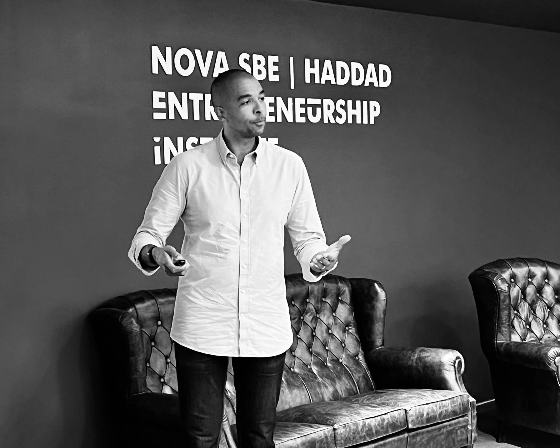 A man in a white shirt giving a presentation in a conference room with leather chairs and a wall with text behind him.