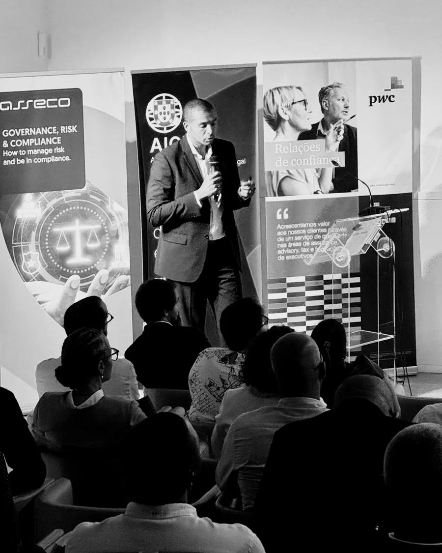 A man giving a presentation at a conference, standing at a podium with a microphone, in front of banners with logos and text. Audience members are seated, facing the speaker.