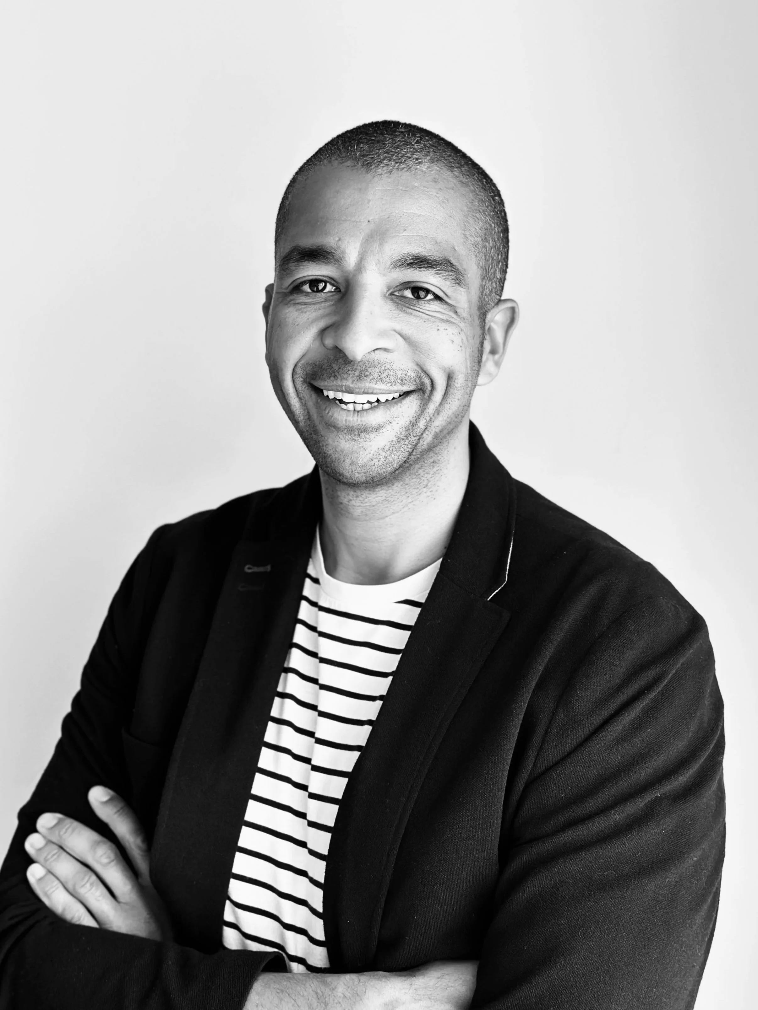 Black and white portrait of a smiling man with short hair, wearing a striped shirt and a dark blazer, standing against a plain light background.