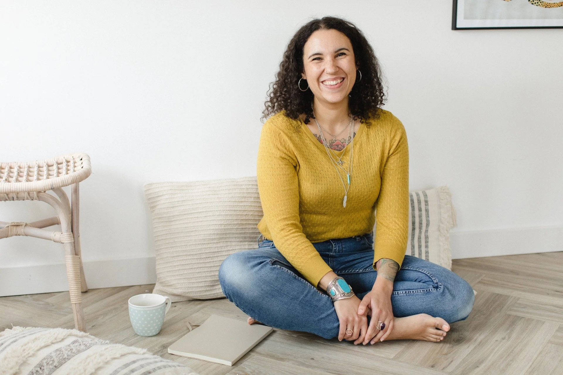 A mixed race woman sitting cross-legged on the floor, smiling, wearing a mustard yellow sweater and blue jeans, with jewelry and tattoos, in a cozy room with a cup, notebook, and a cushion around her.
