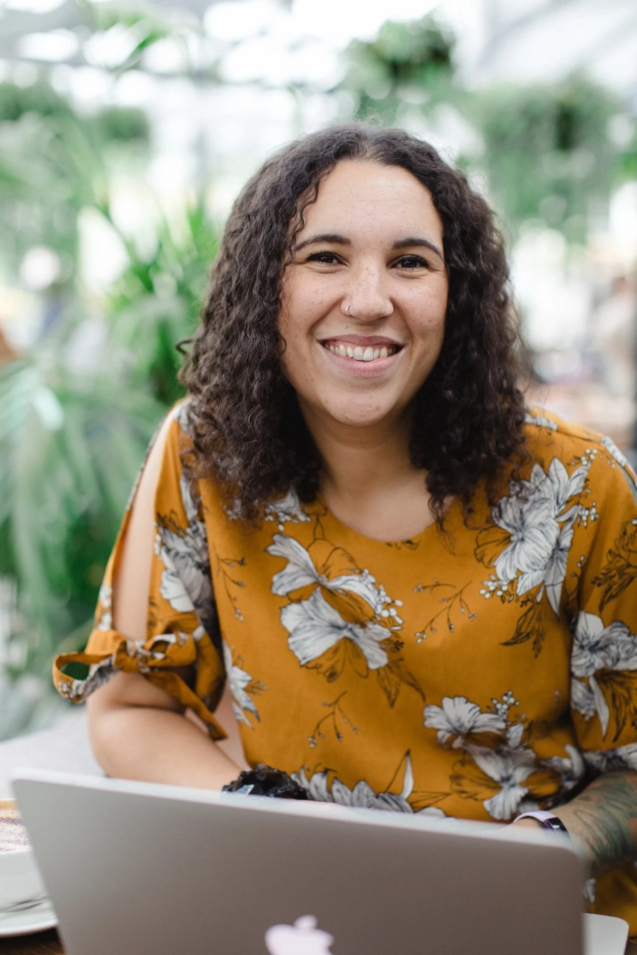 A woman with curly dark hair, wearing a yellow floral top, smiling at the camera while sitting at a table with a laptop in a bright, green indoor setting.