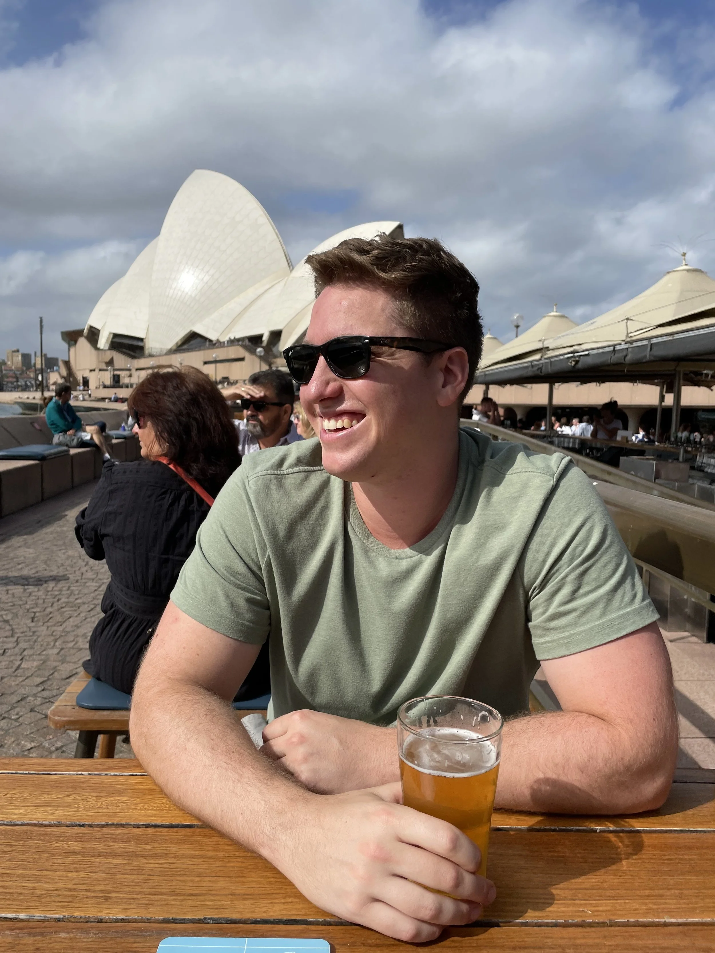 A young man in sunglasses sitting outdoors at a wooden table with a glass of beer in front of him, smiling. The Sydney Opera House is visible in the background under a partly cloudy sky.