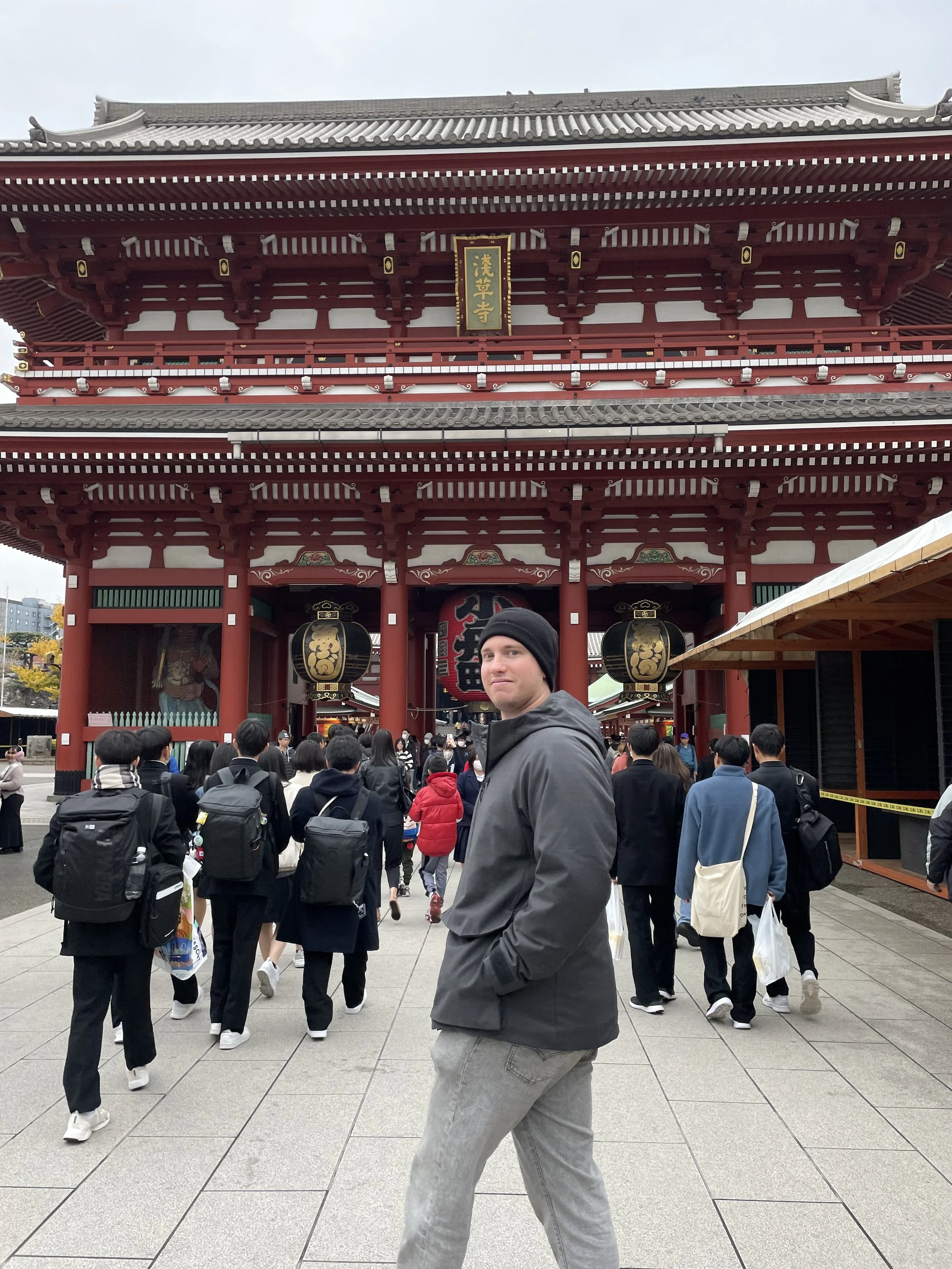 A man in a black jacket and gray pants posing in front of a traditional Japanese temple entrance with a large lantern and many people walking under and around it.