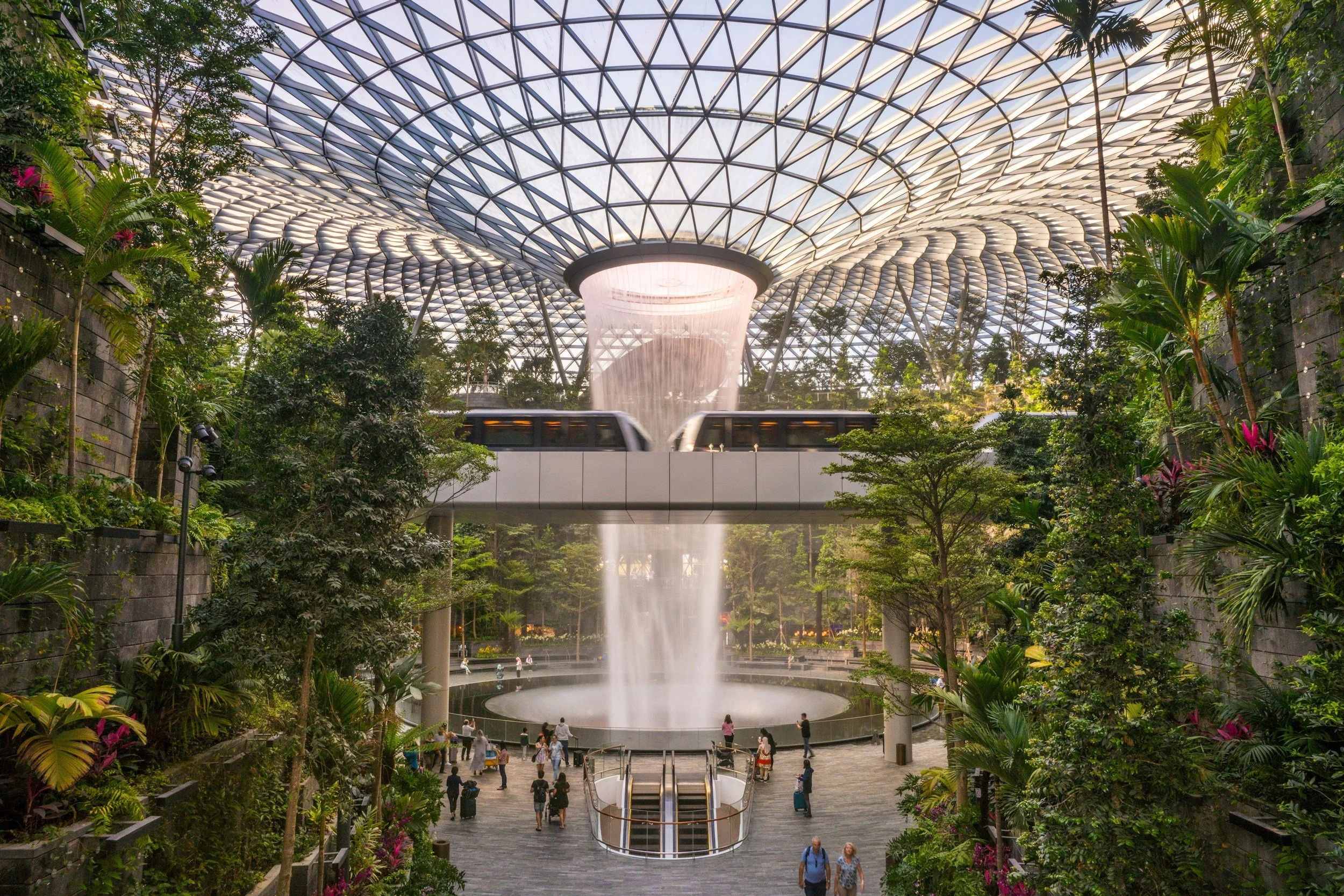 Interior of a large indoor garden with lush green plants, a waterfall feature, and a glass geometric dome ceiling. A monorail train passes over an open area with visitors walking below.