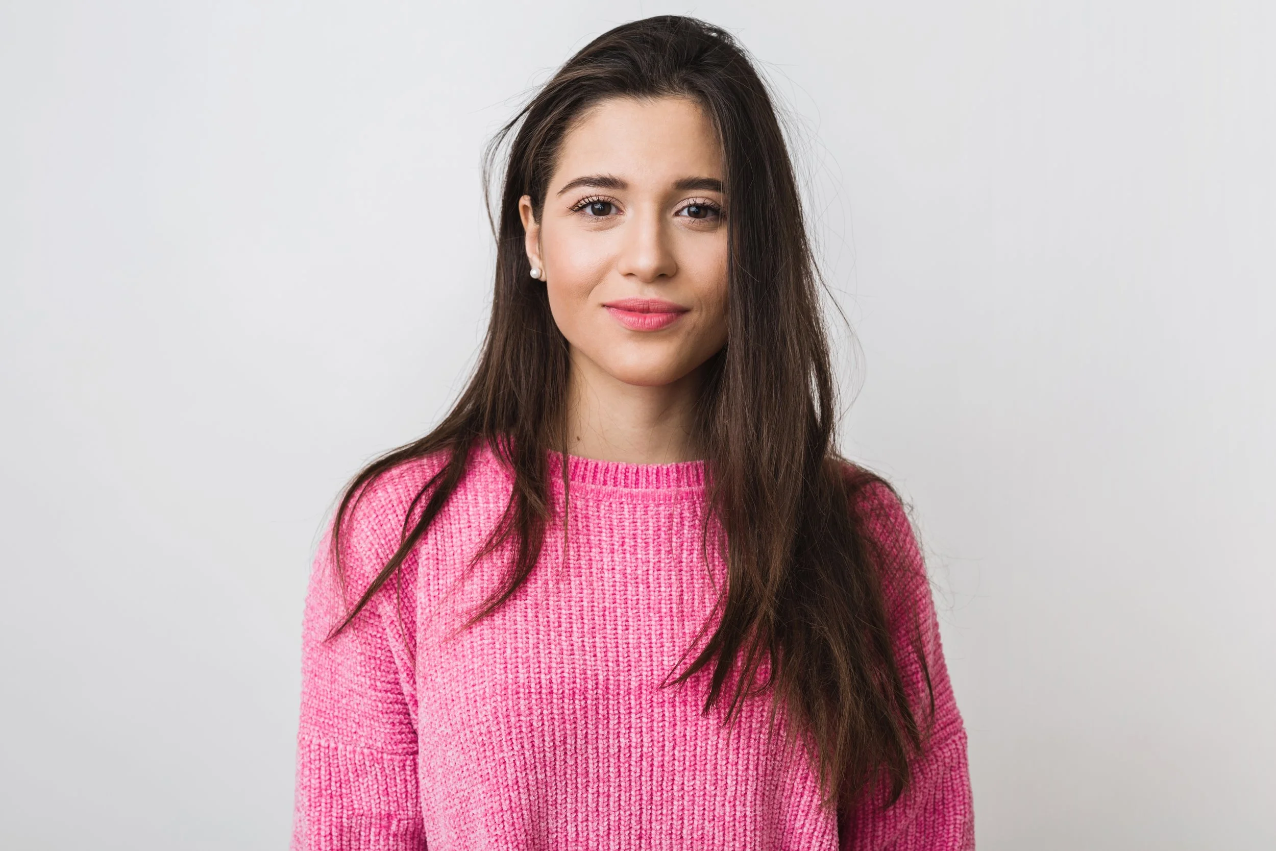 A young woman with long brown hair, wearing a pink sweater, standing against a plain white background.