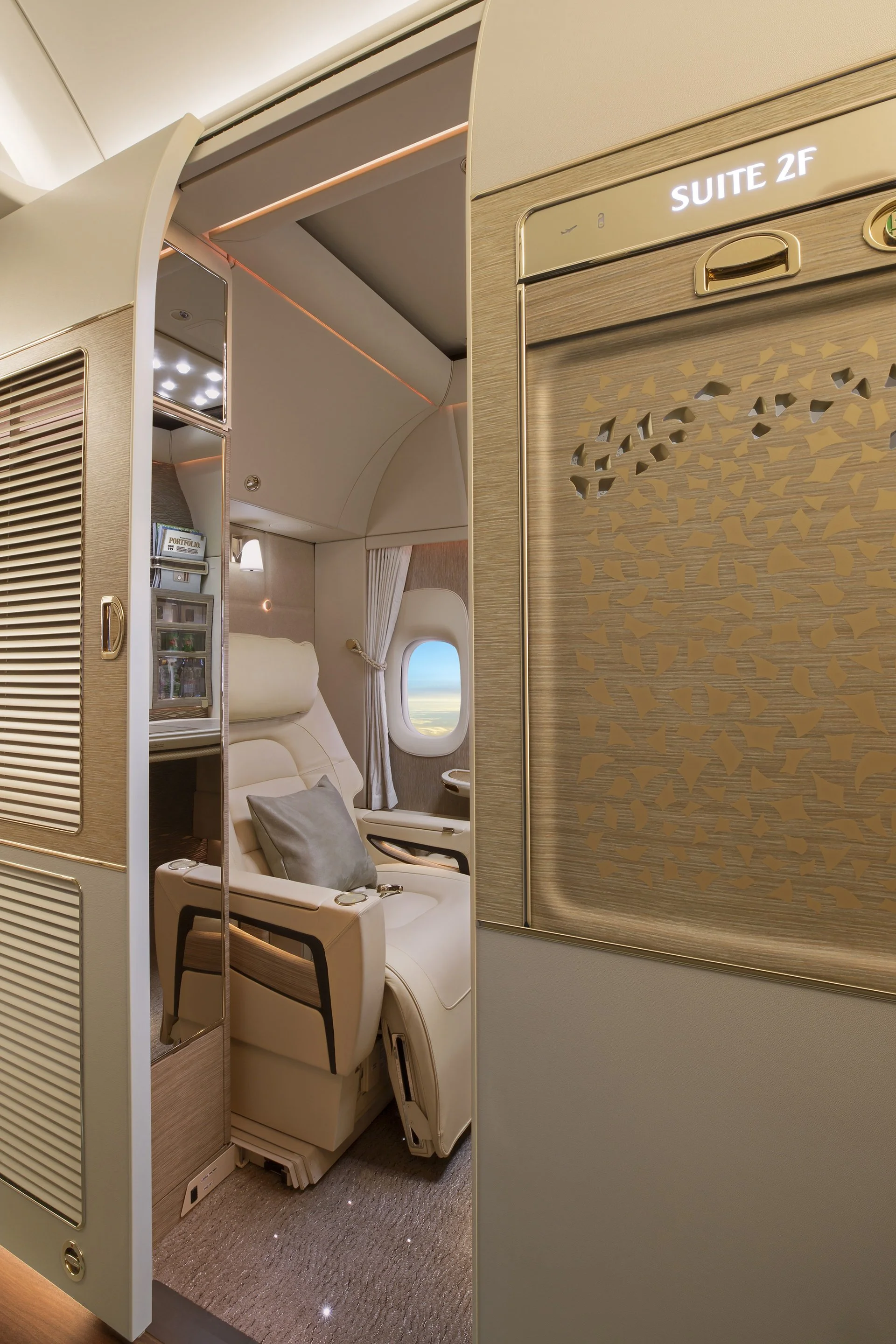 Inside a first class airplane cabin, showing a luxurious lie-flat seat with a pillow, nearby window with a view of the sky, and gold accents with the label 'Suite 2F' on the wall.