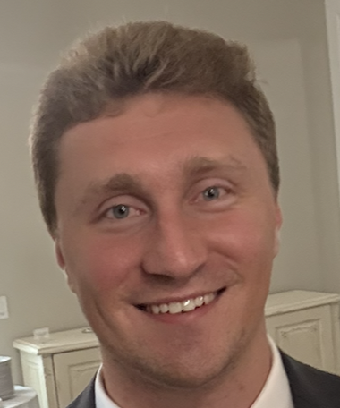 A young man with dark hair, glasses, and a light beard smiling outdoors in front of snowy trees, wearing a black hoodie.