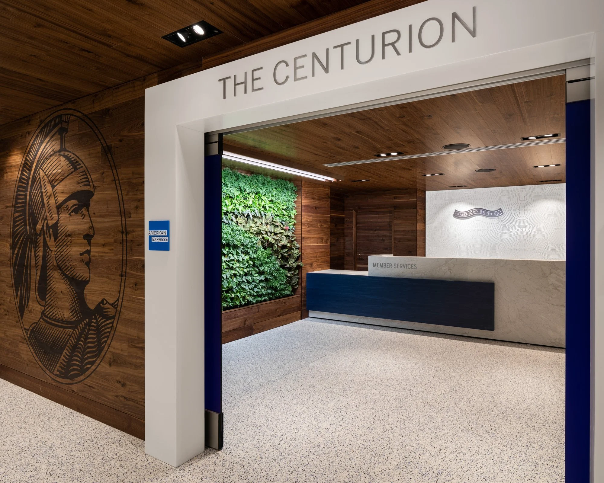 Entrance to a lobby area with a sign reading 'The Centurion' and a reception desk labeled 'Member Services.' The walls feature the American Express logo, with a lush green vertical garden and a large graphic of a Native American chief in profile on the side wall. Modern lighting and a wood-paneled ceiling create a warm, inviting atmosphere.
